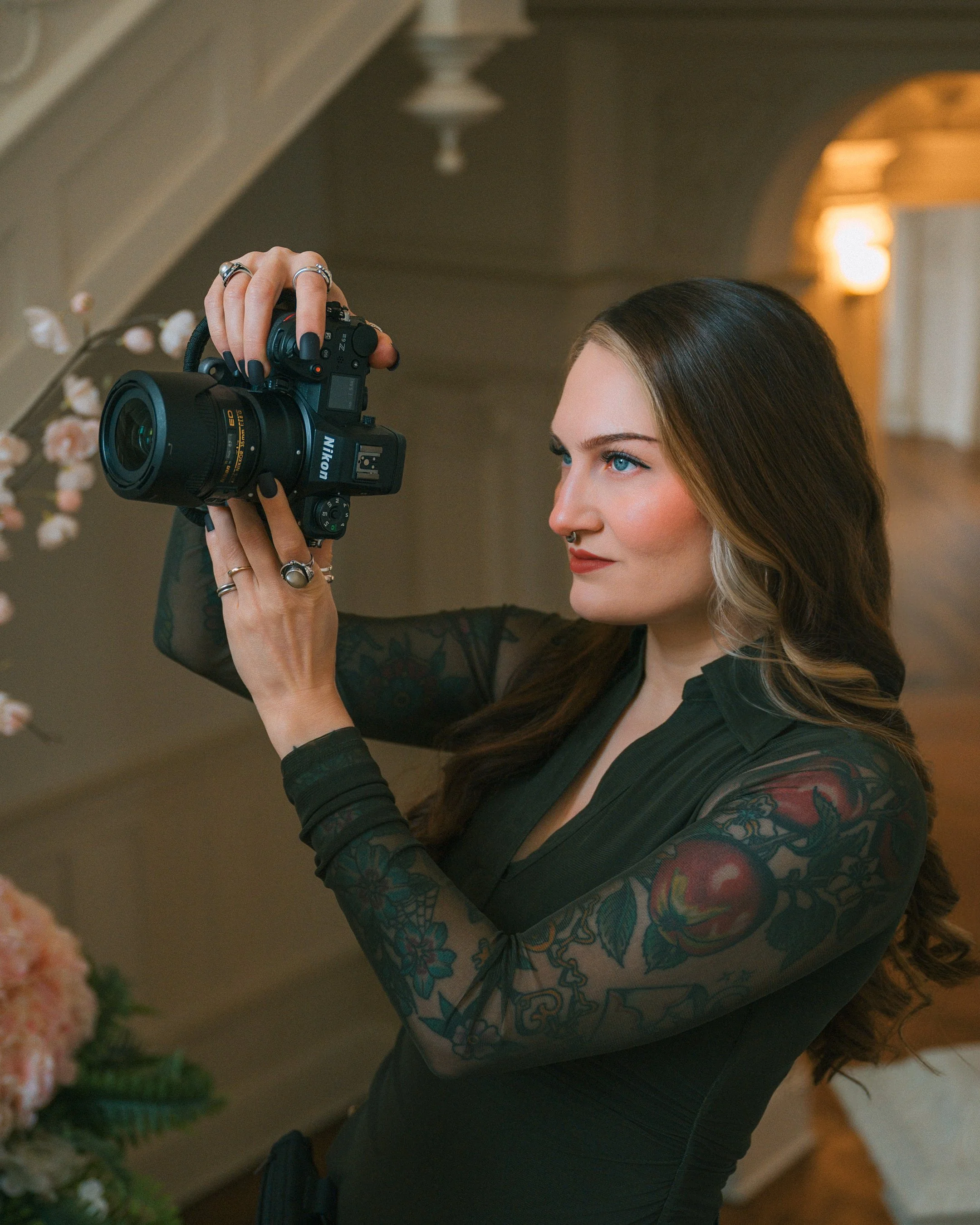 A woman with long brown hair and blue eyes taking a photo with a Nikon camera, wearing a black dress with floral lace sleeves, inside a house with soft lighting and wooden decor.