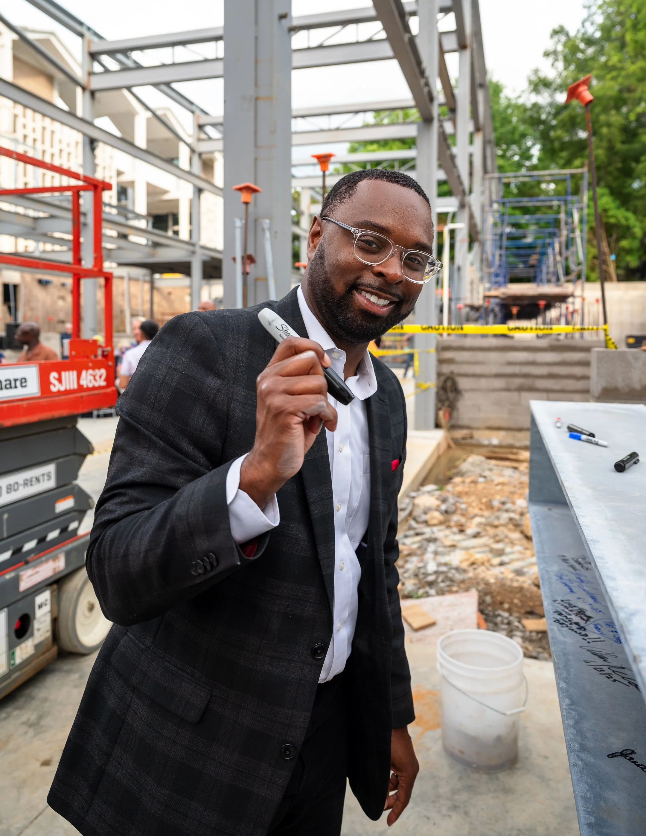 A man in a suit holding a marker at a construction site, smiling at the camera.