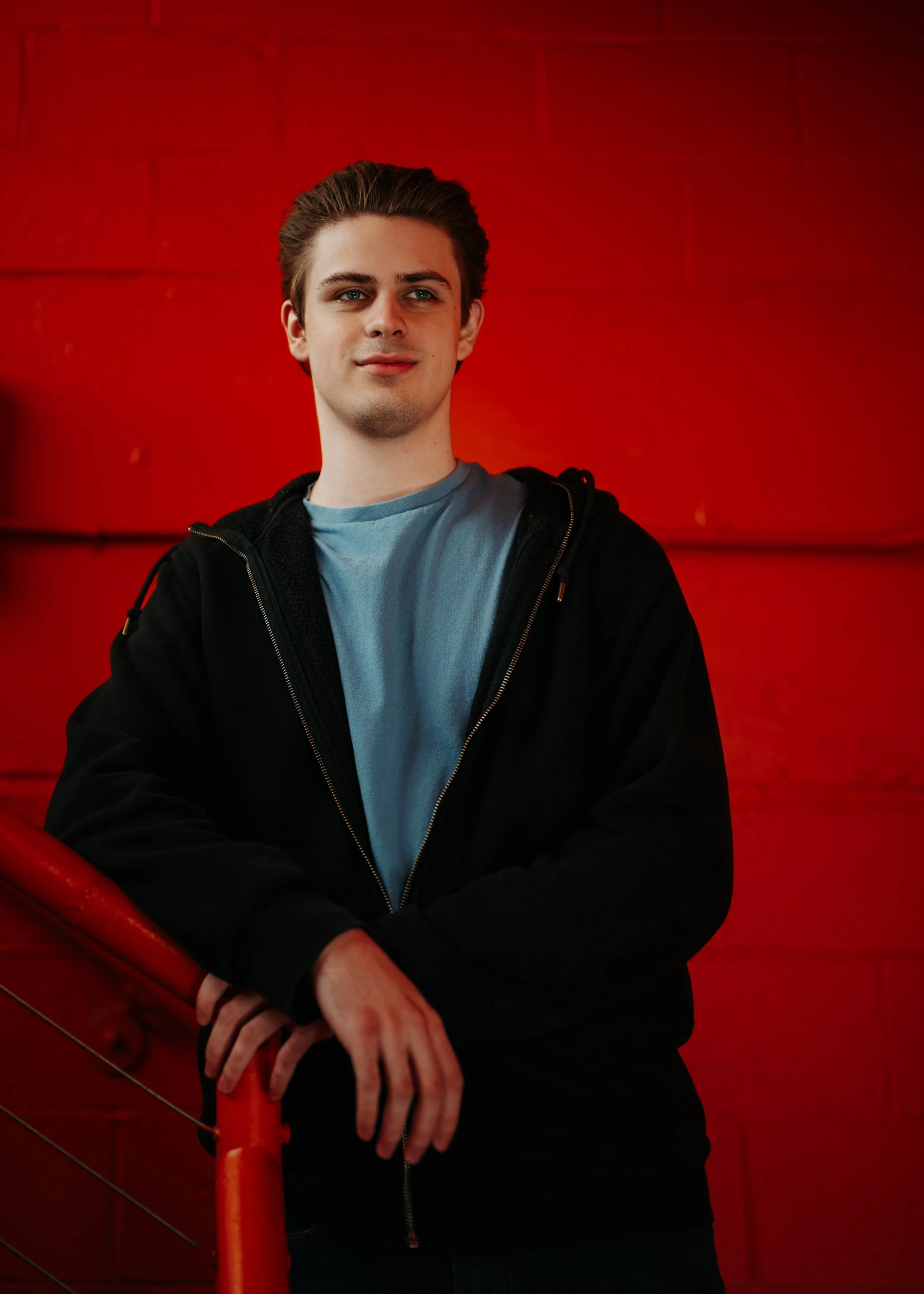 A young man with brown hair, blue eyes, and light skin, wearing a black hoodie over a light blue t-shirt, standing against a red wall. He is holding onto a red railing with his left hand and has a slight smile.