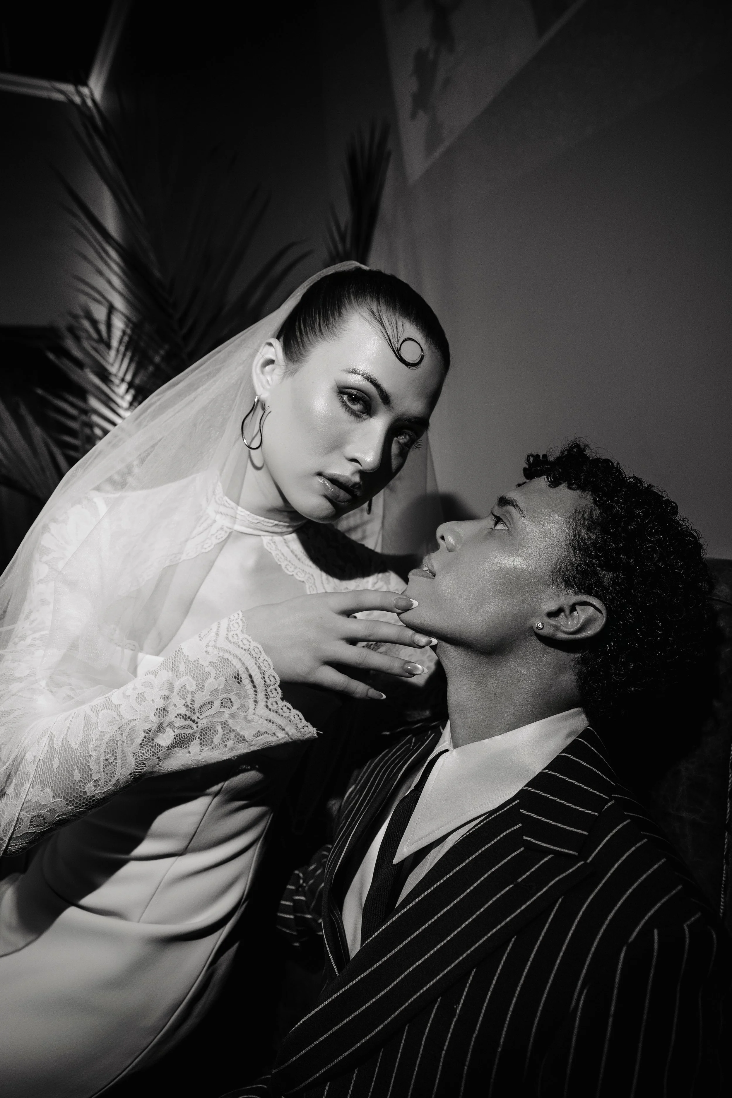 A black-and-white photograph of two women, one lying down and the other leaning over her. The woman lying down has short, curly hair, and wears a striped jacket. The woman leaning over has long hair styled in a tight bun, wears earrings, and a lace v