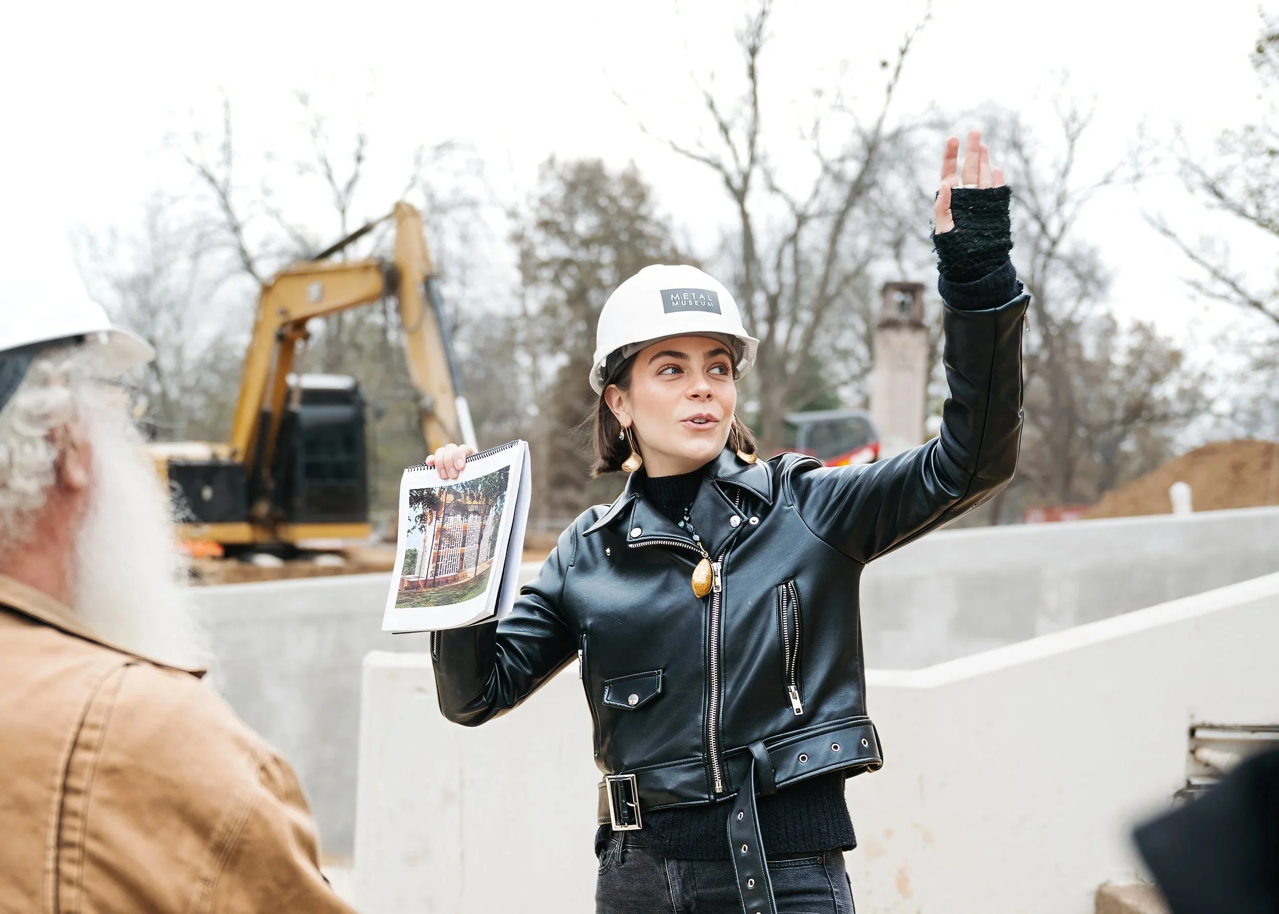 Construction site with a woman wearing a safety helmet and black leather jacket, holding a photo and speaking to a man with a beard in a brown jacket, with an excavator in the background.