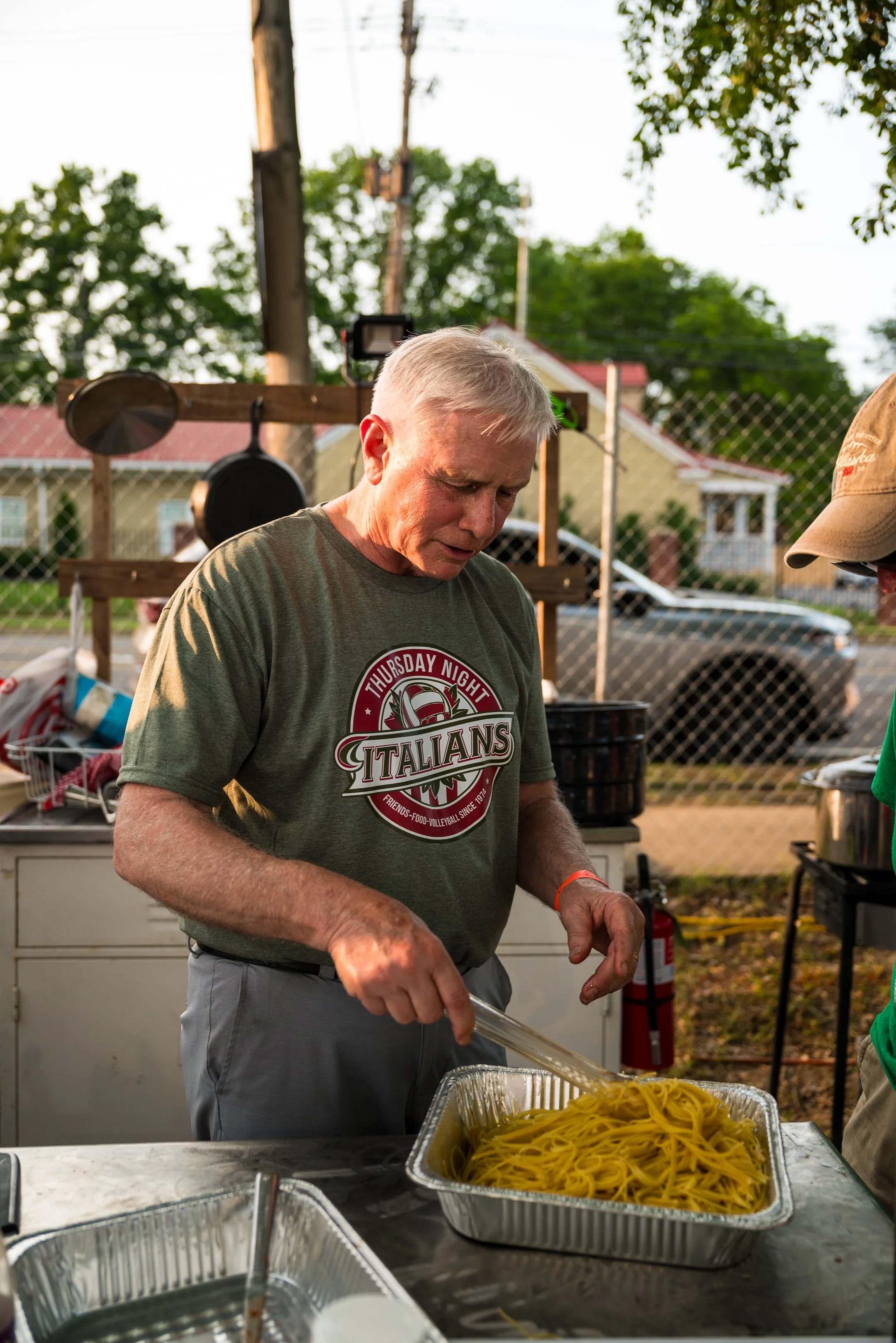 An elderly man with gray hair wearing a green T-shirt and gray pants is serving cooked spaghetti pasta from a large aluminum tray at an outdoor event during daylight. There are cooking utensils hanging in the background, and a chain-link fence with t