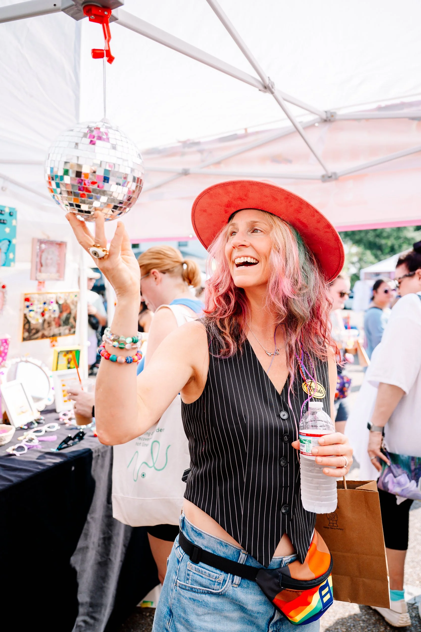 A woman with pink and green hair wearing a red hat, a sleeveless pinstripe shirt, and blue jeans, smiling and spinning a small disco ball hanging from a tent ceiling at an outdoor event.