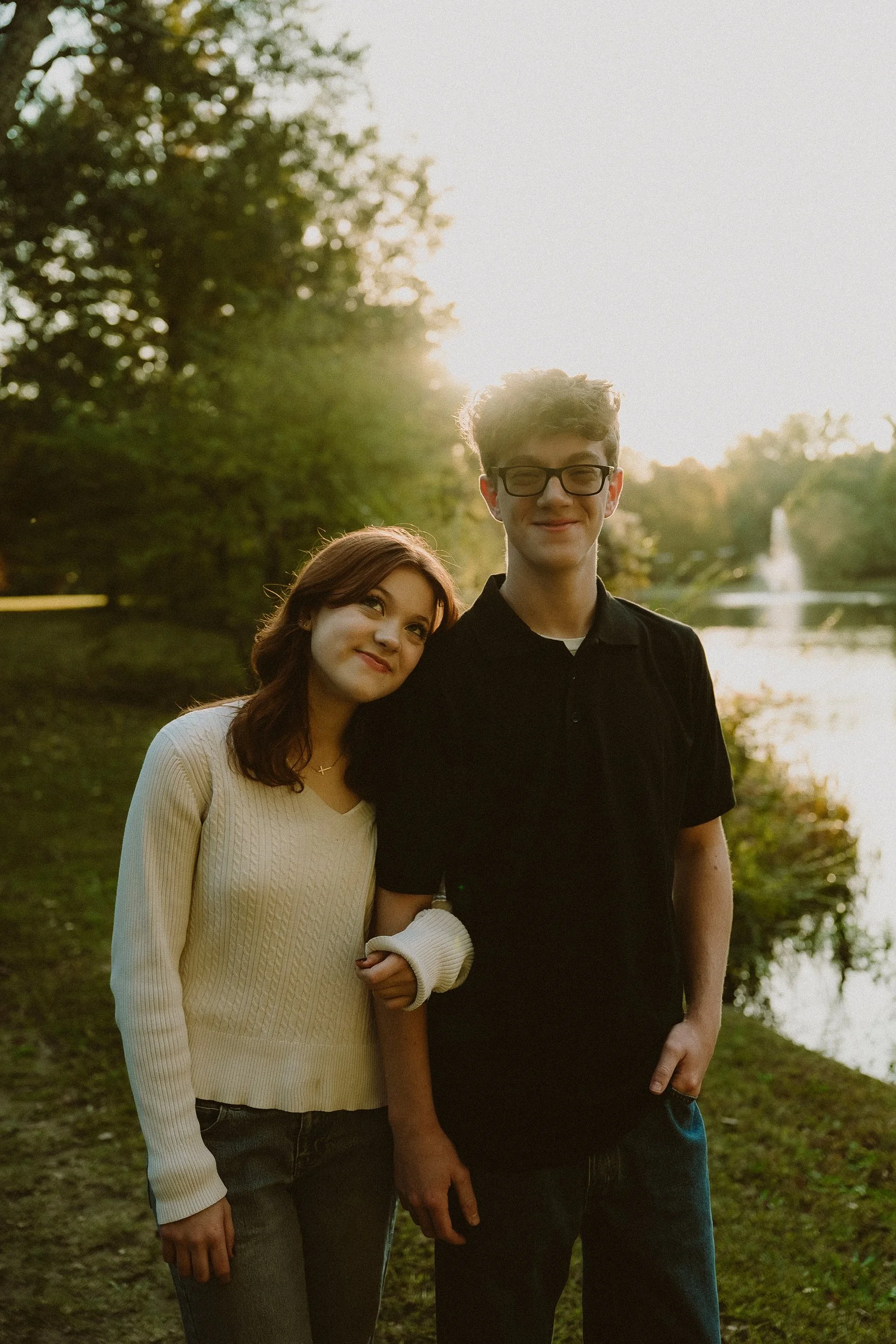 A young woman and young man standing together outdoors by a lake during sunset, with trees and a fountain in the background.
