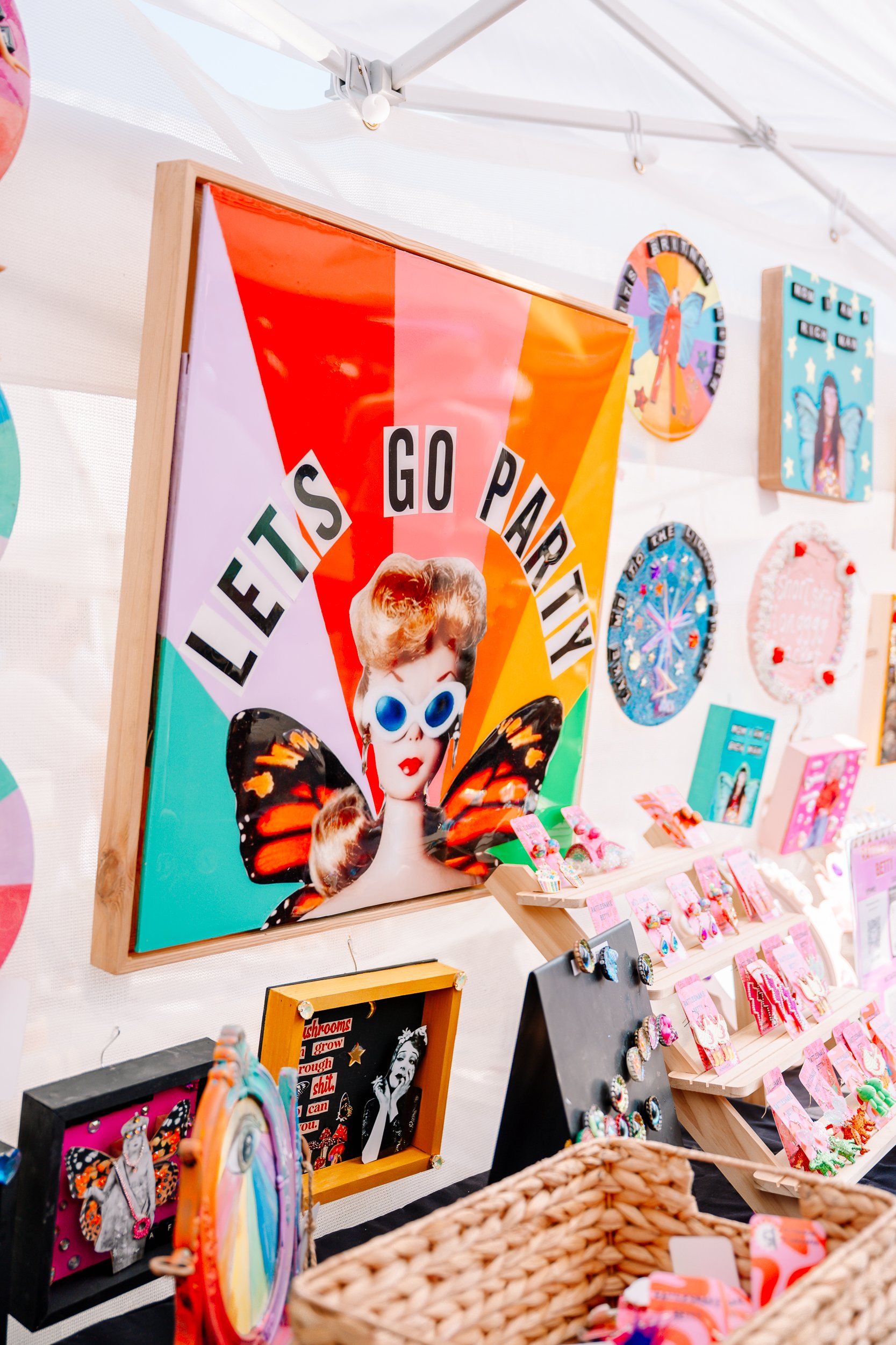 Colorful display at a marketplace or craft fair featuring a sign with a collage of a woman with butterfly wings and the text "Let's Go Party", surrounded by various art pieces and jewelry.