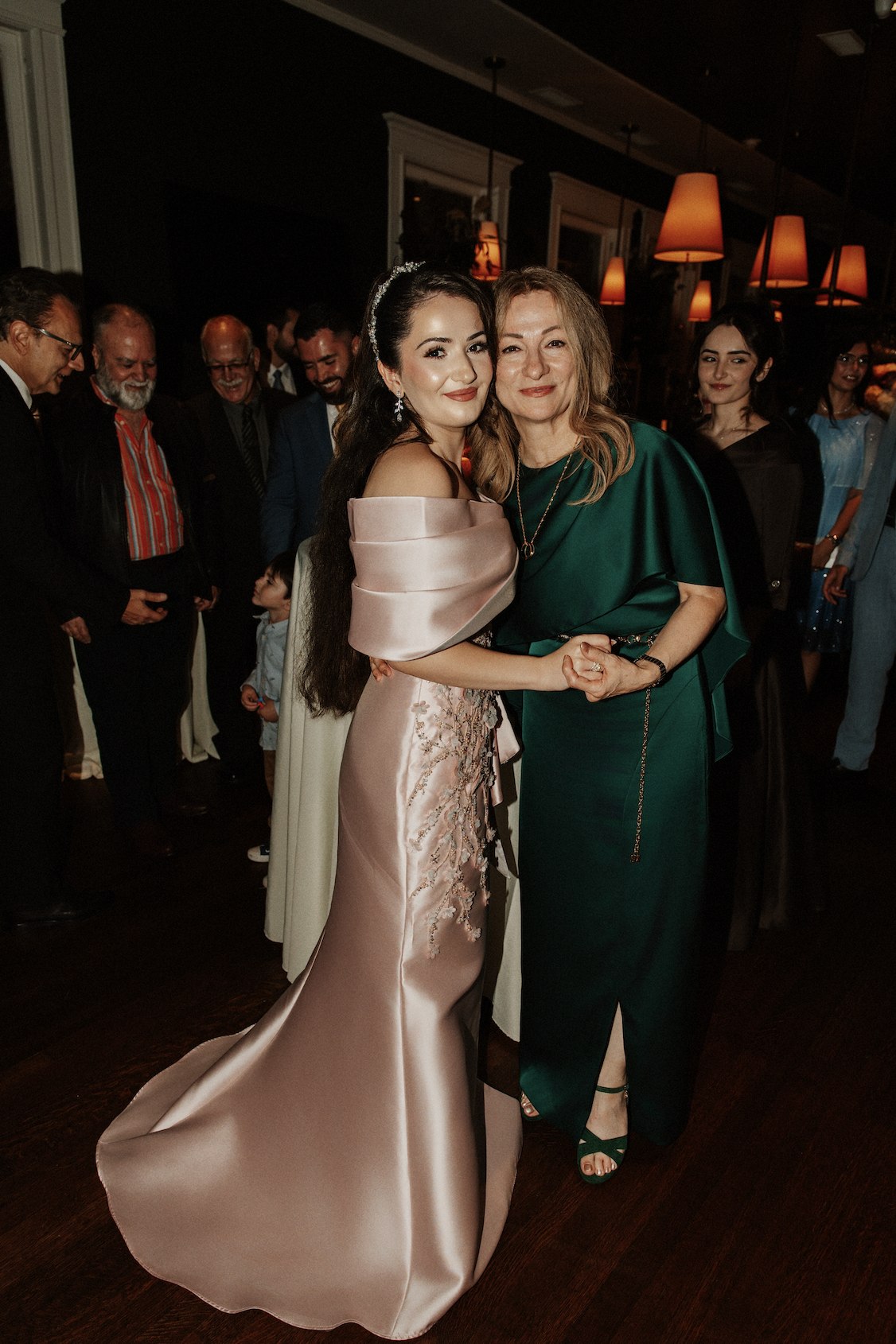 A young woman in a pink satin gown stands beside an older woman in a green dress, both smiling and holding hands at a social event with other people in the background.