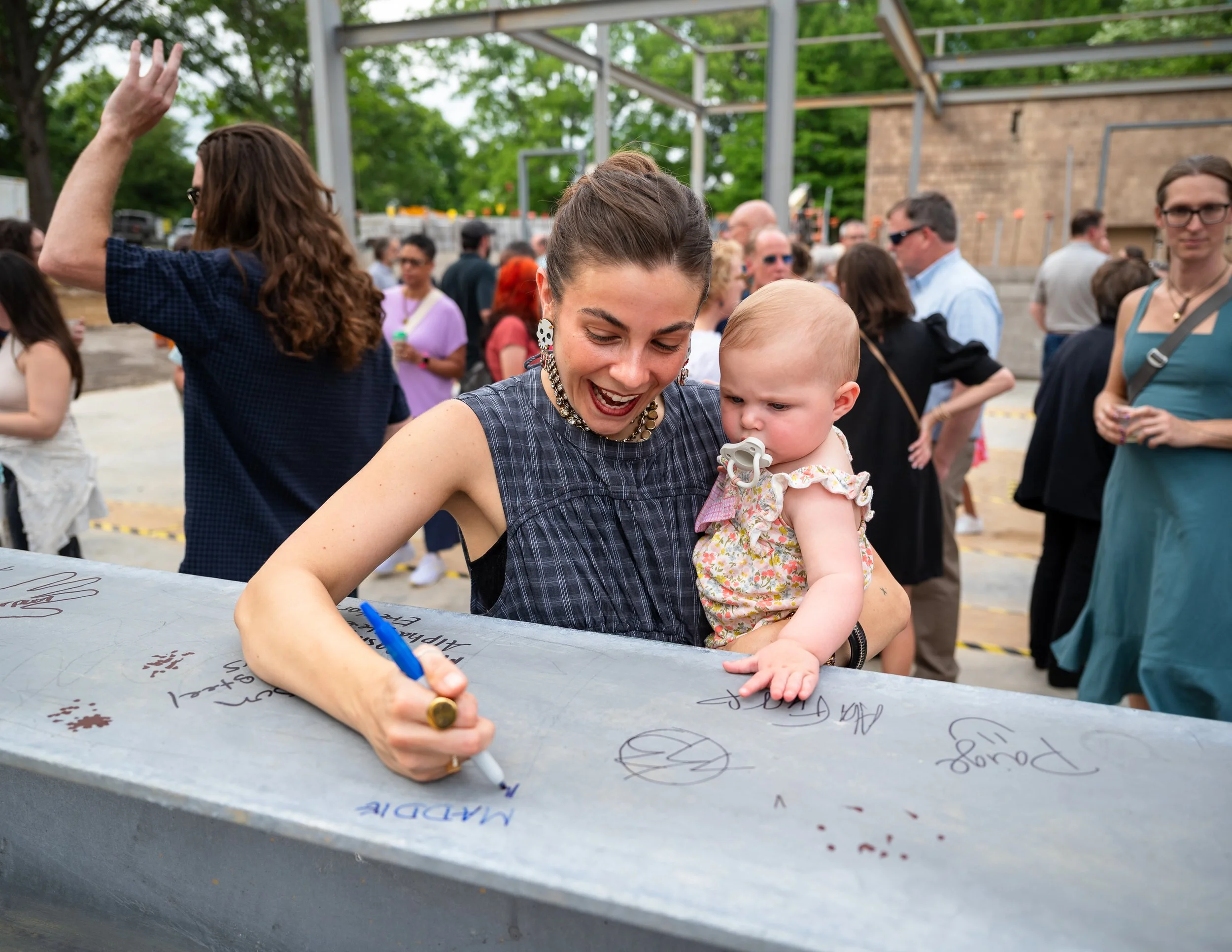 A woman with dark hair and earrings signs a metal surface with a blue marker, while a young child with a pacifier leans on her shoulder. Several people are gathered in the background at an outdoor event.