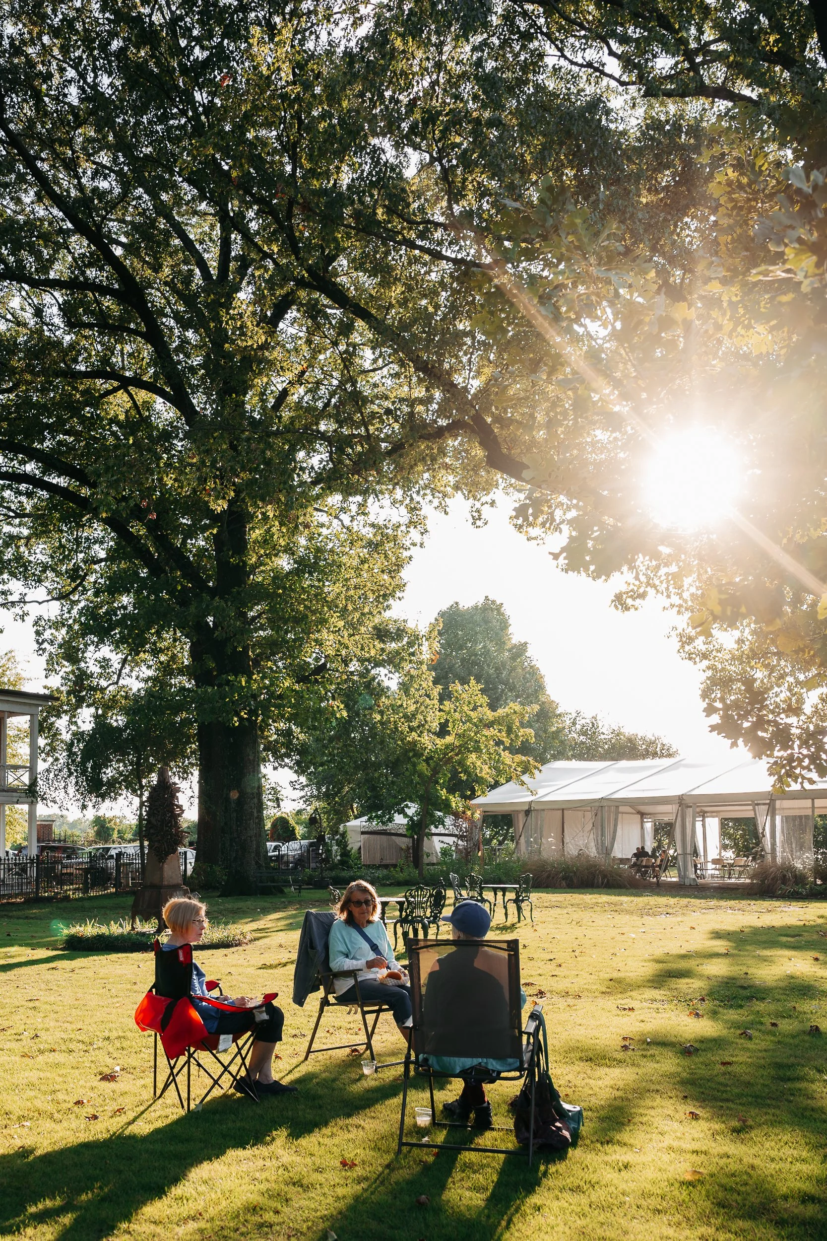 People sitting outdoors on folding chairs in a garden area with large trees and a bright sun in the sky, near a large white tent.