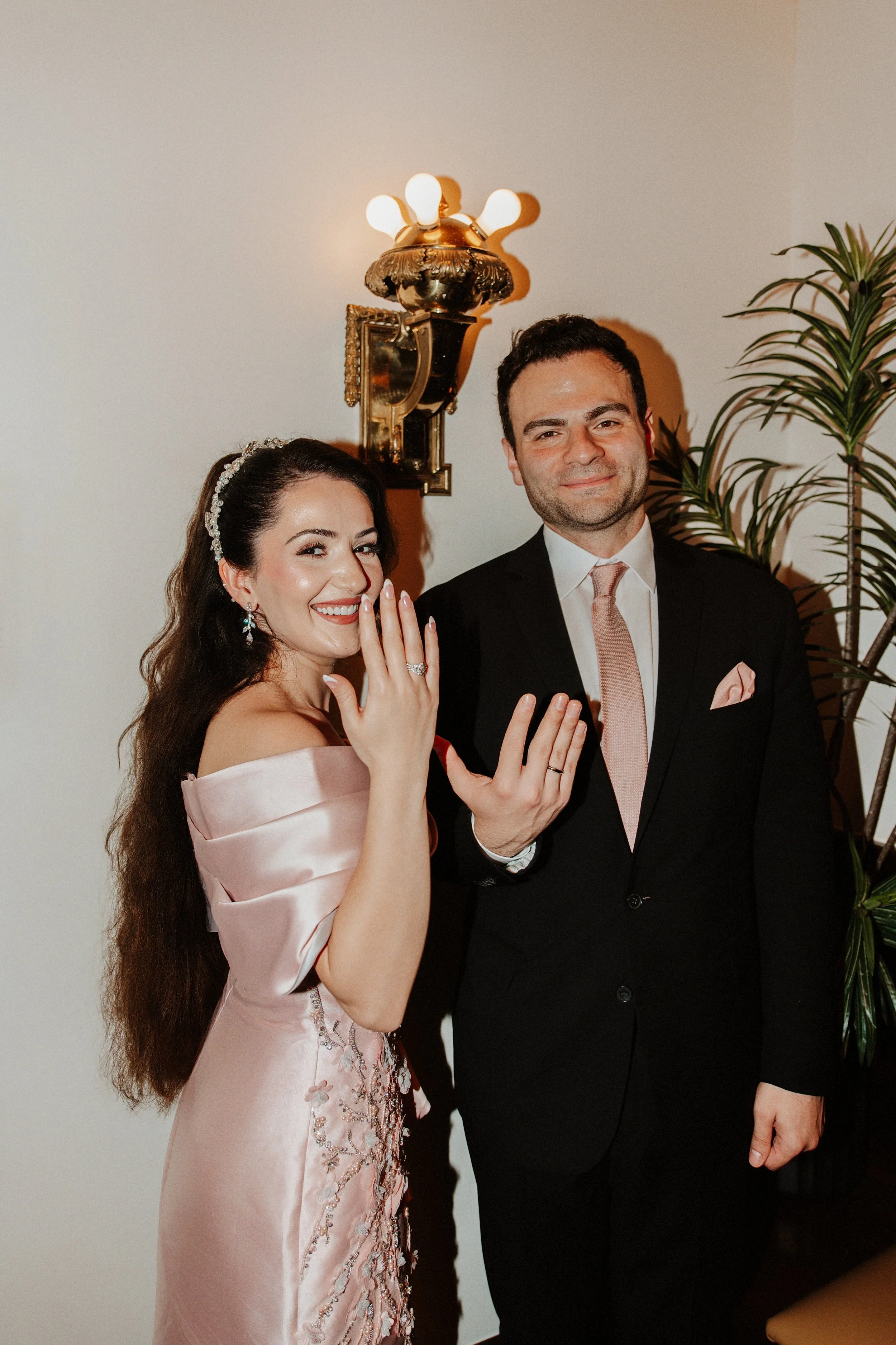 A bride and groom showing off their wedding rings, smiling at the camera, indoors with a white wall, a decorative wall lamp, and a potted plant in the background.