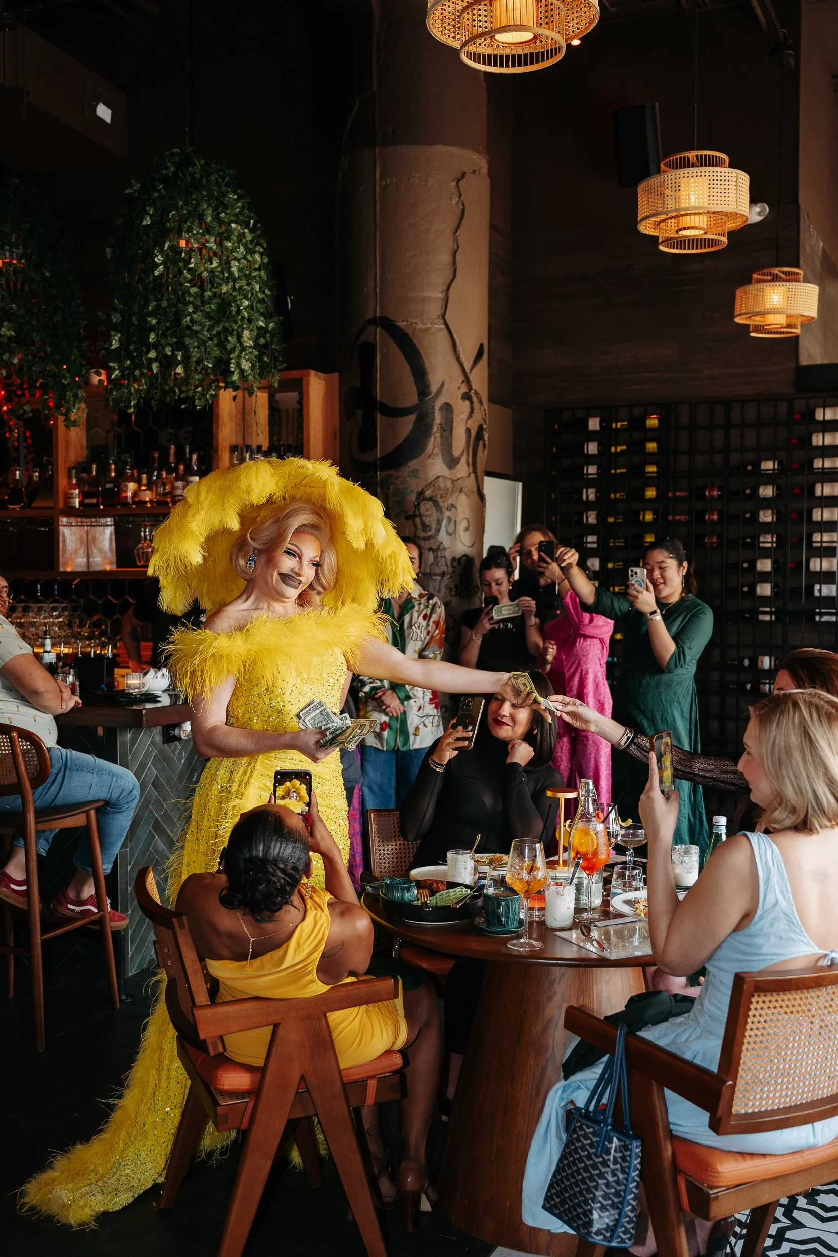 A drag queen in a bright yellow feathered gown performing a money dance for guests seated at a restaurant table, with others taking photos and videos.