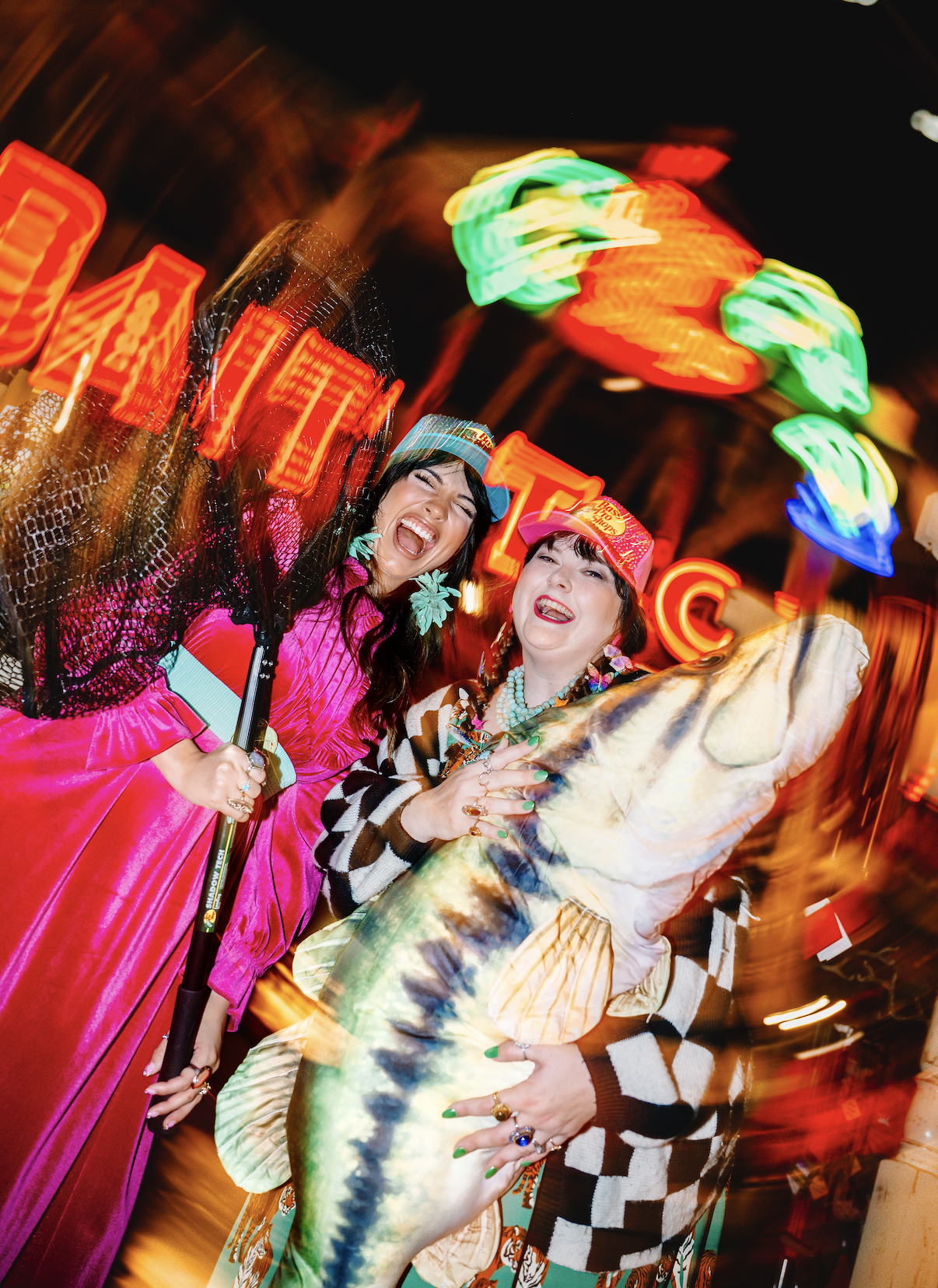 Two women dressed in colorful, playful clothing holding large fish toy, laughing and posing in front of a brightly lit amusement park ride at night.