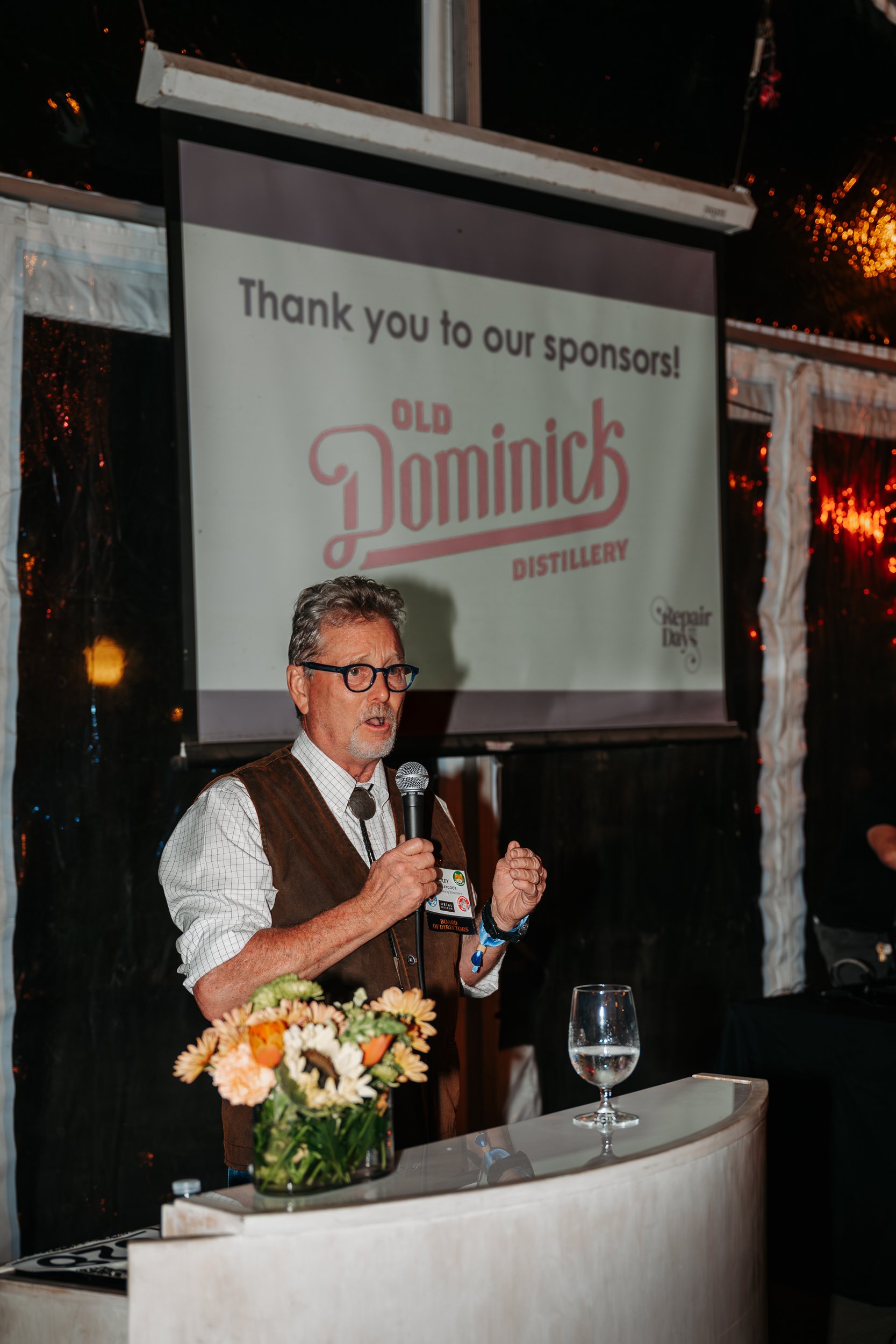 A man in glasses and a vest speaking into a microphone at an event. There is a floral arrangement on the podium and a glass of water. A screen behind him displays a message thanking sponsors, with the logo of Old Dominick Distillery.