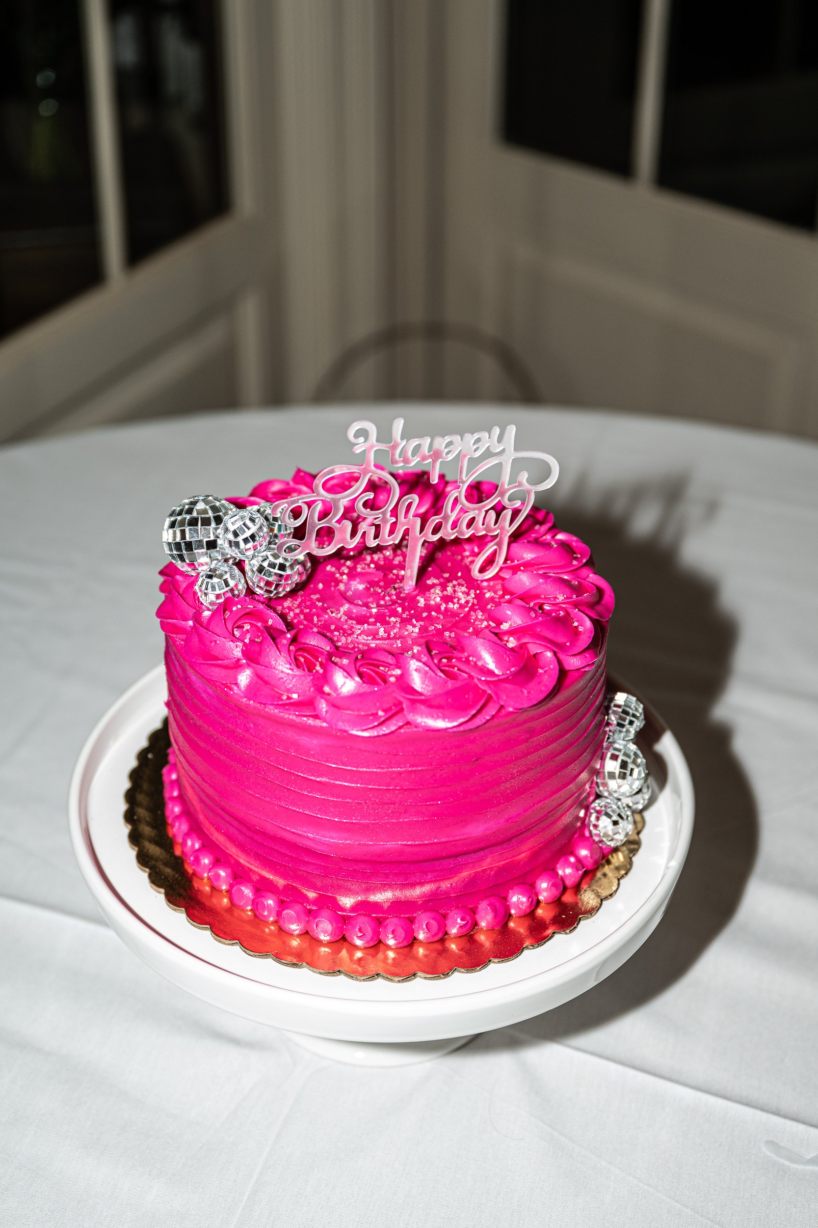 A bright pink birthday cake decorated with silver disco ball ornaments, featuring a 'Happy Birthday' topper, pearl-like embellishments, and swirl piping on top, placed on a white cake stand.
