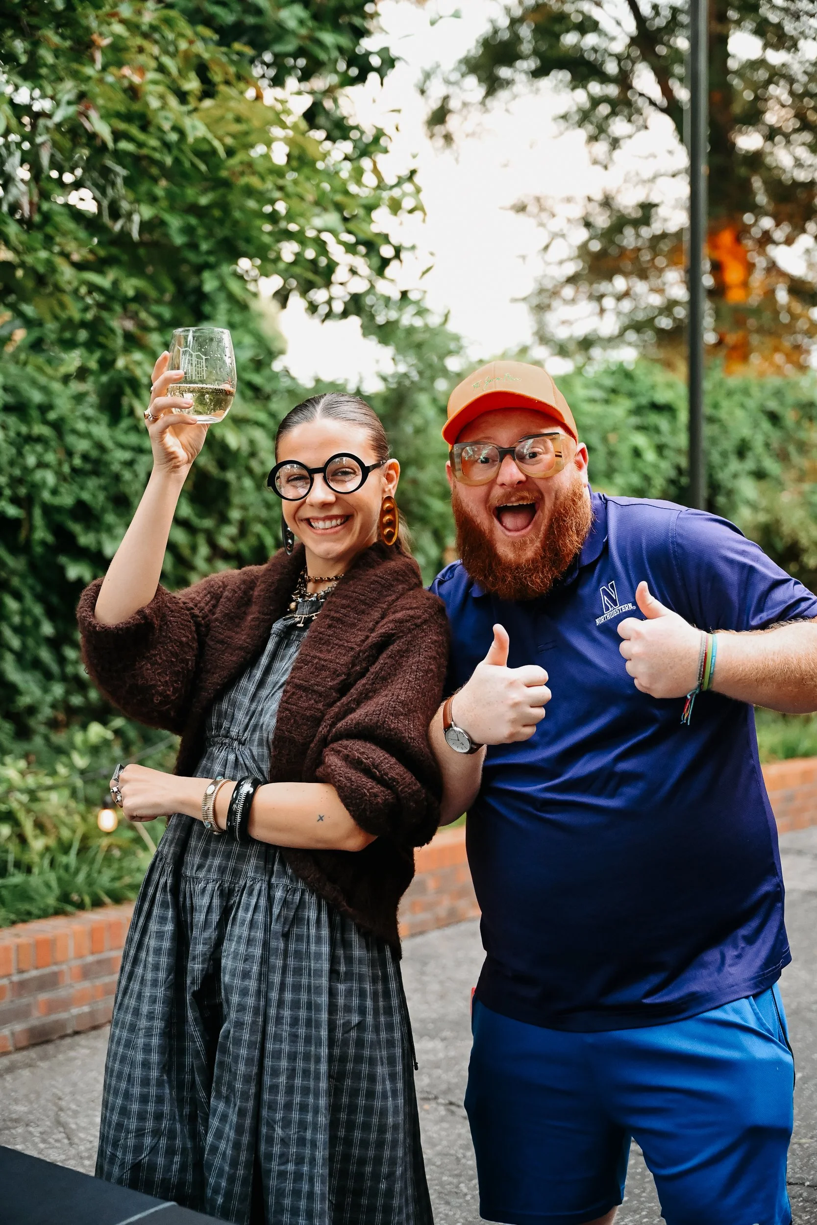Two friends outdoors, smiling and celebrating, one holding a wine glass, the other giving a thumbs-up.