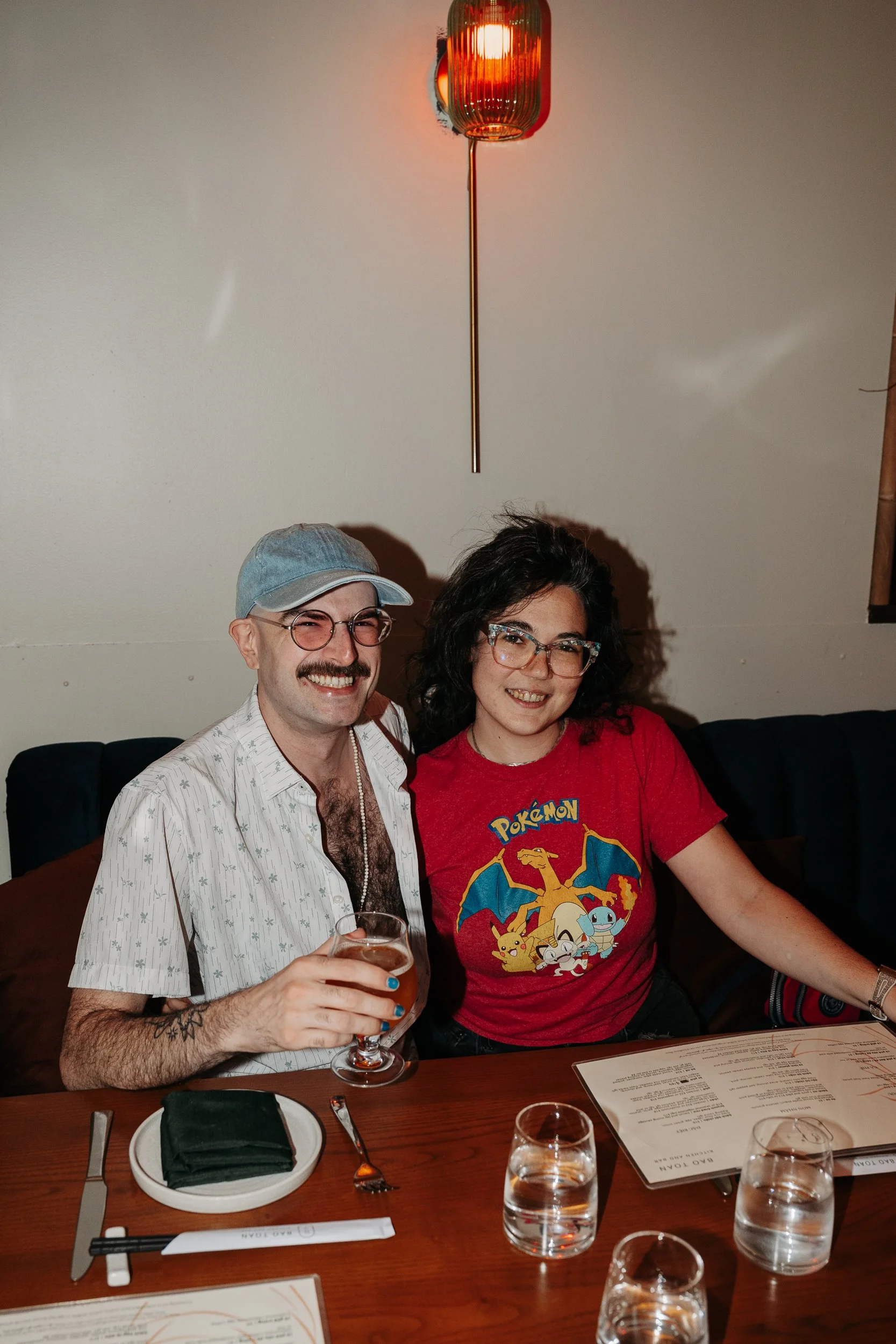 Two people sitting at a restaurant table, smiling at the camera. The person on the left is a man with glasses, a mustache, wearing a denim cap and a short-sleeved shirt. The person on the right is a woman with curly dark hair, glasses, wearing a red 