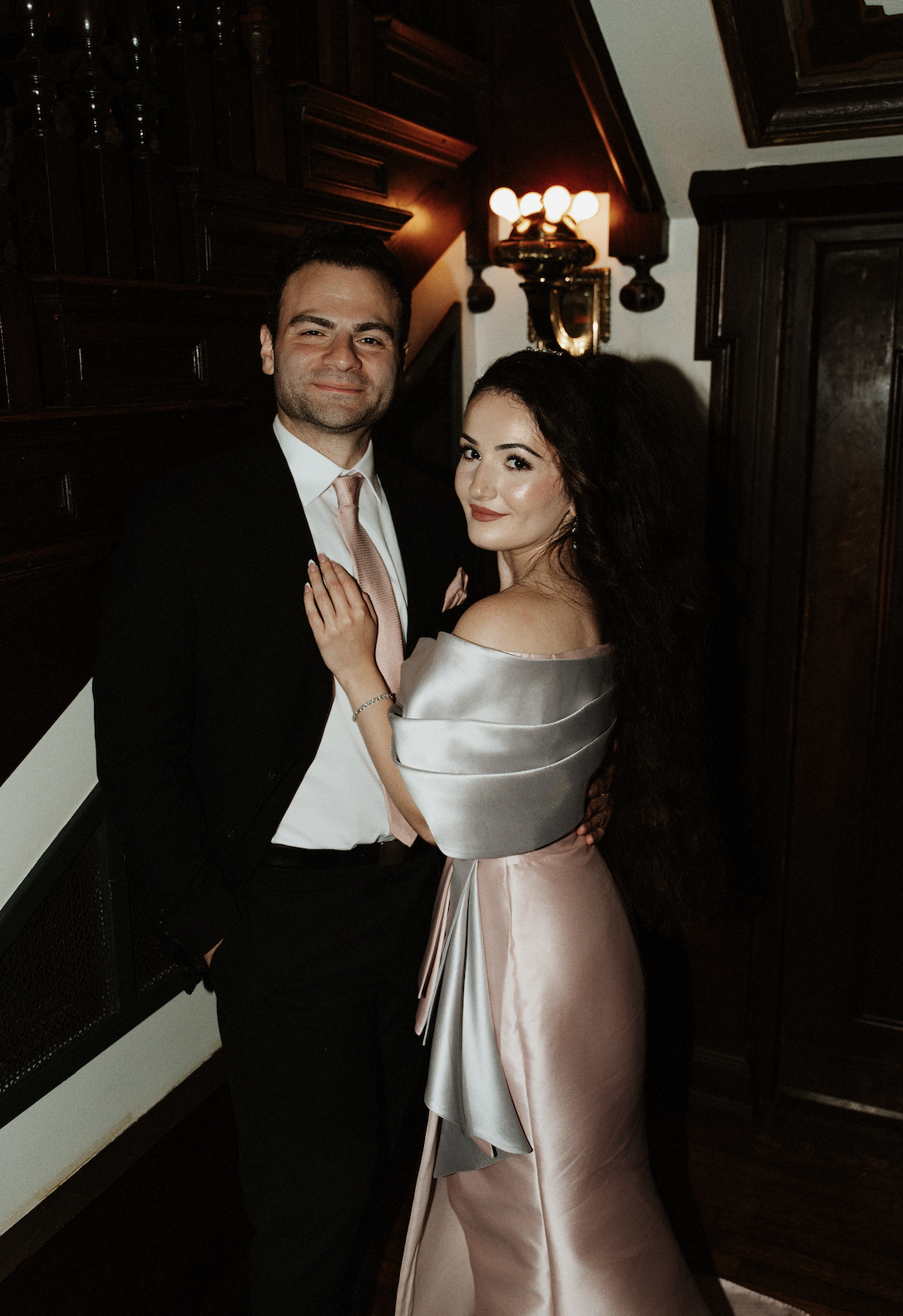 A man and woman dressed in formal attire, posing together indoors with dark wood paneling and antique lighting in the background.