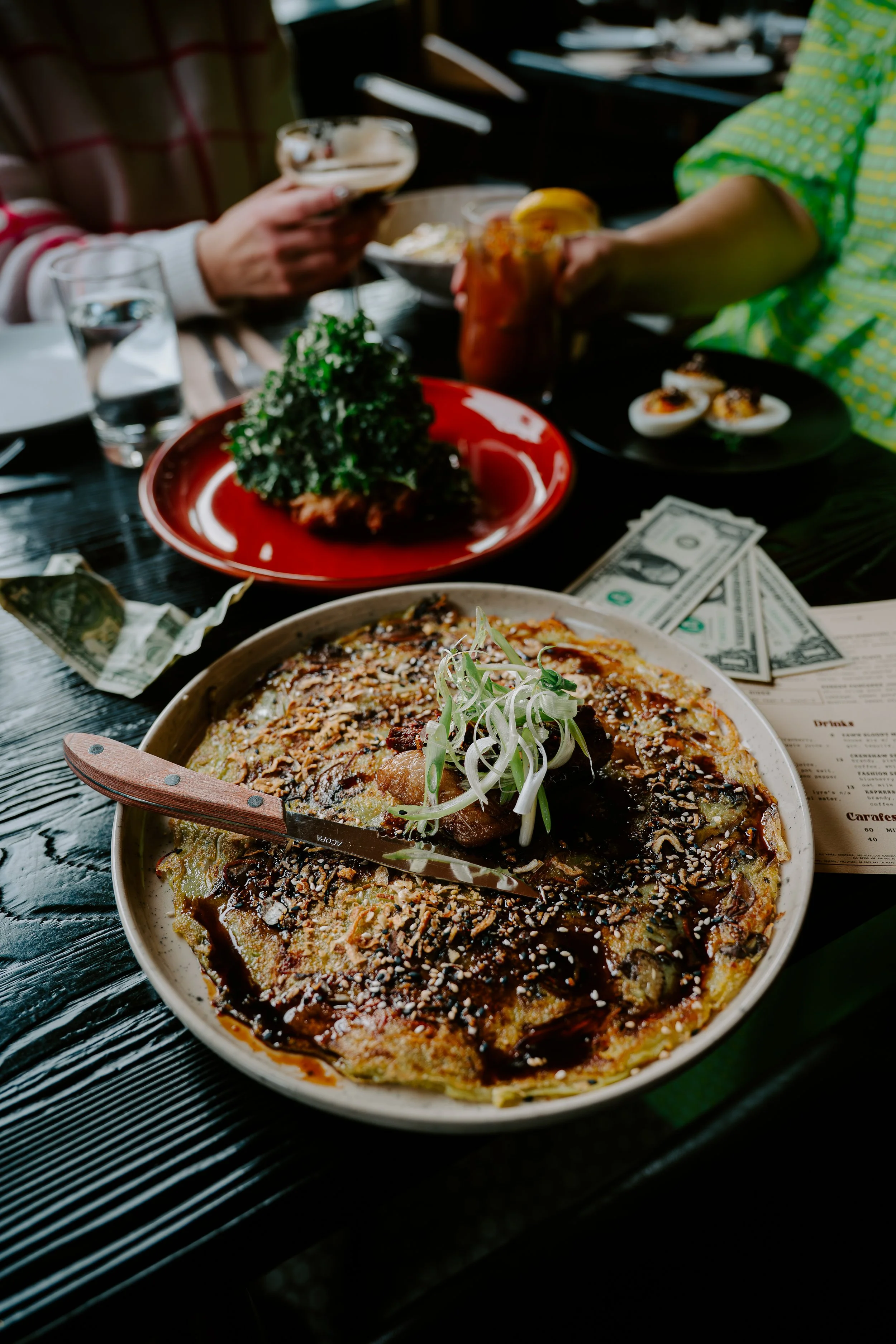 Close-up of a pizza with vegetables and sesame seeds, on a black wooden table. In the background, there is a partially eaten dish, some drinks, and money on the table.