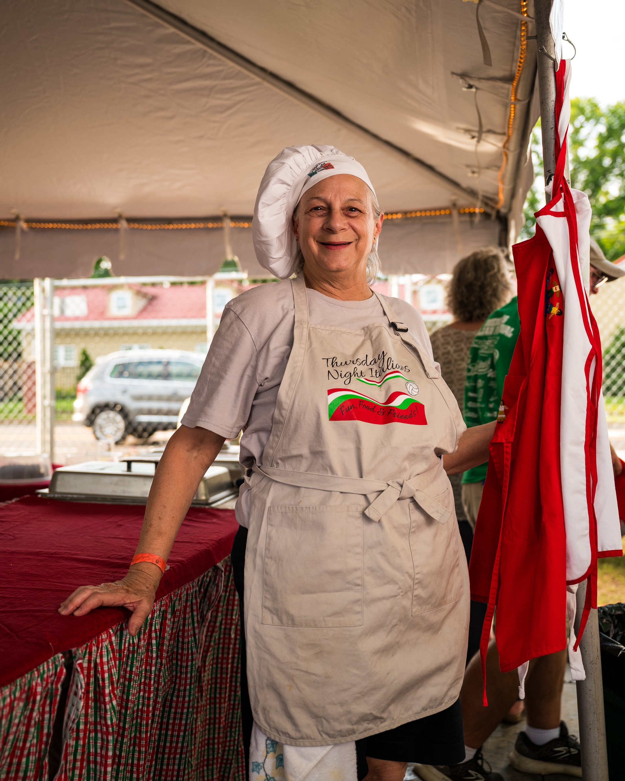 Smiling woman wearing a chef's hat and apron standing at a food stand at an outdoor event.