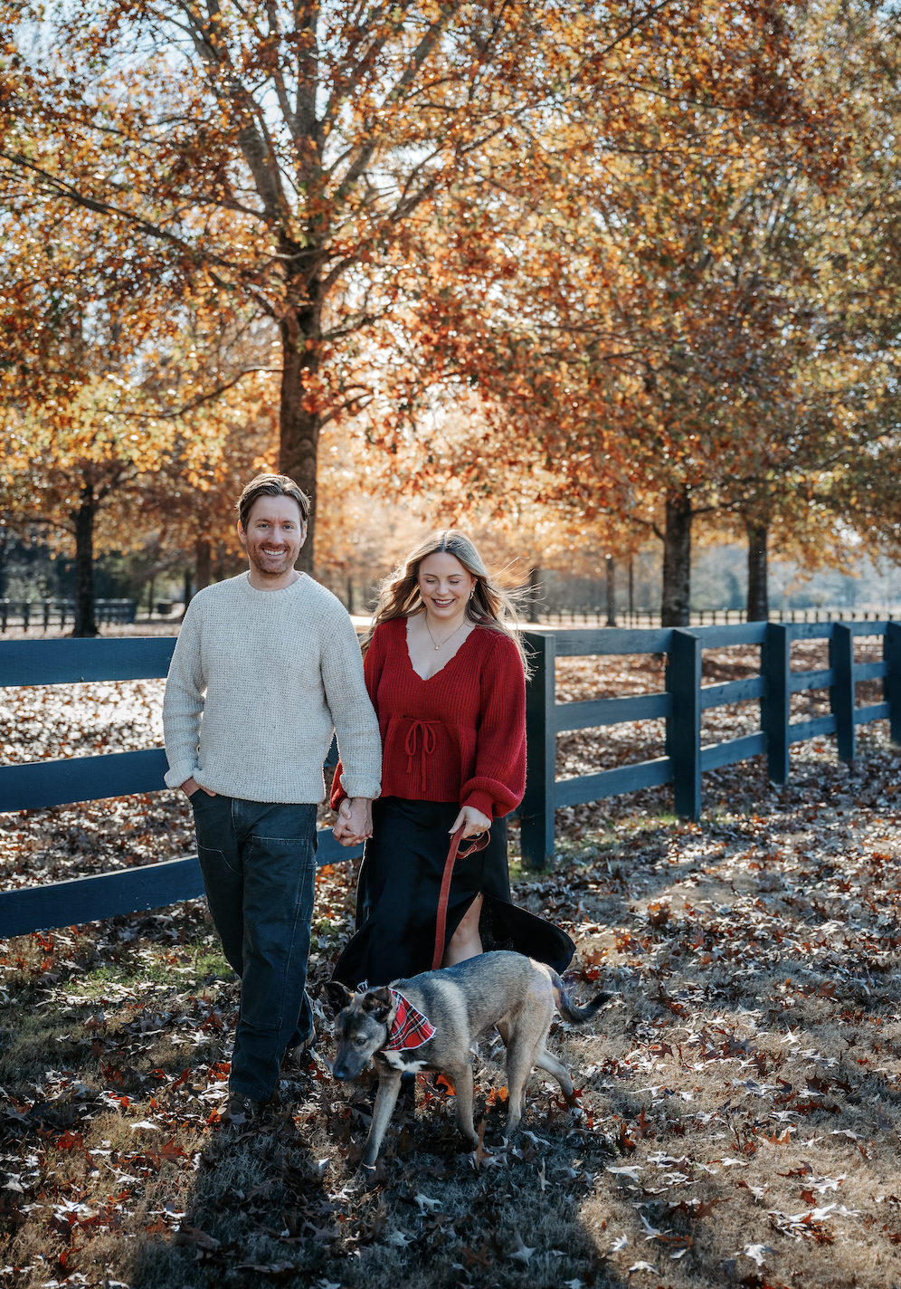 A happy couple walking hand-in-hand with their dog in a park during fall, surrounded by orange and brown autumn leaves, trees, and a wooden fence.