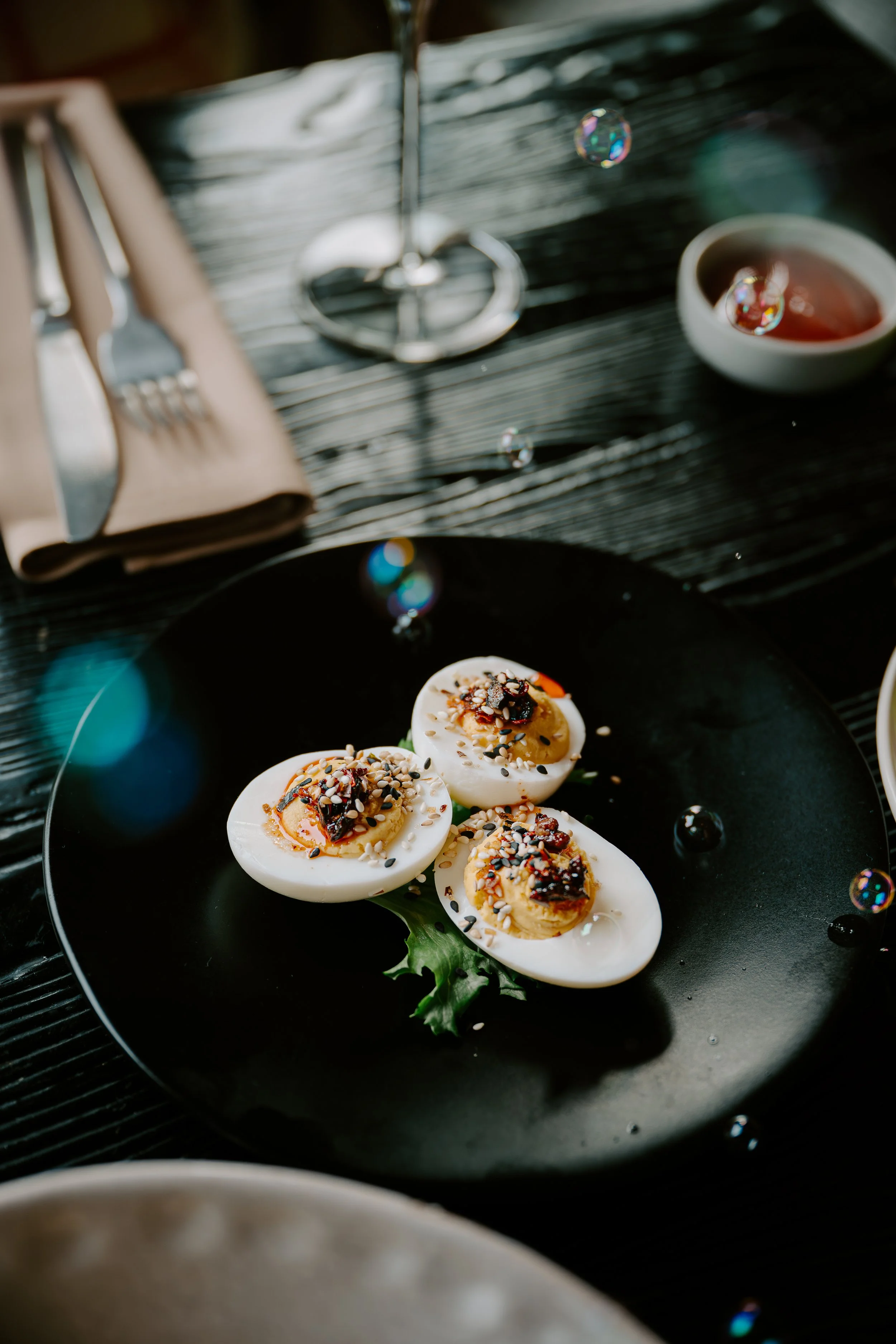 A black plate with halved boiled eggs topped with seasoning and sauce, placed on a dark textured table.