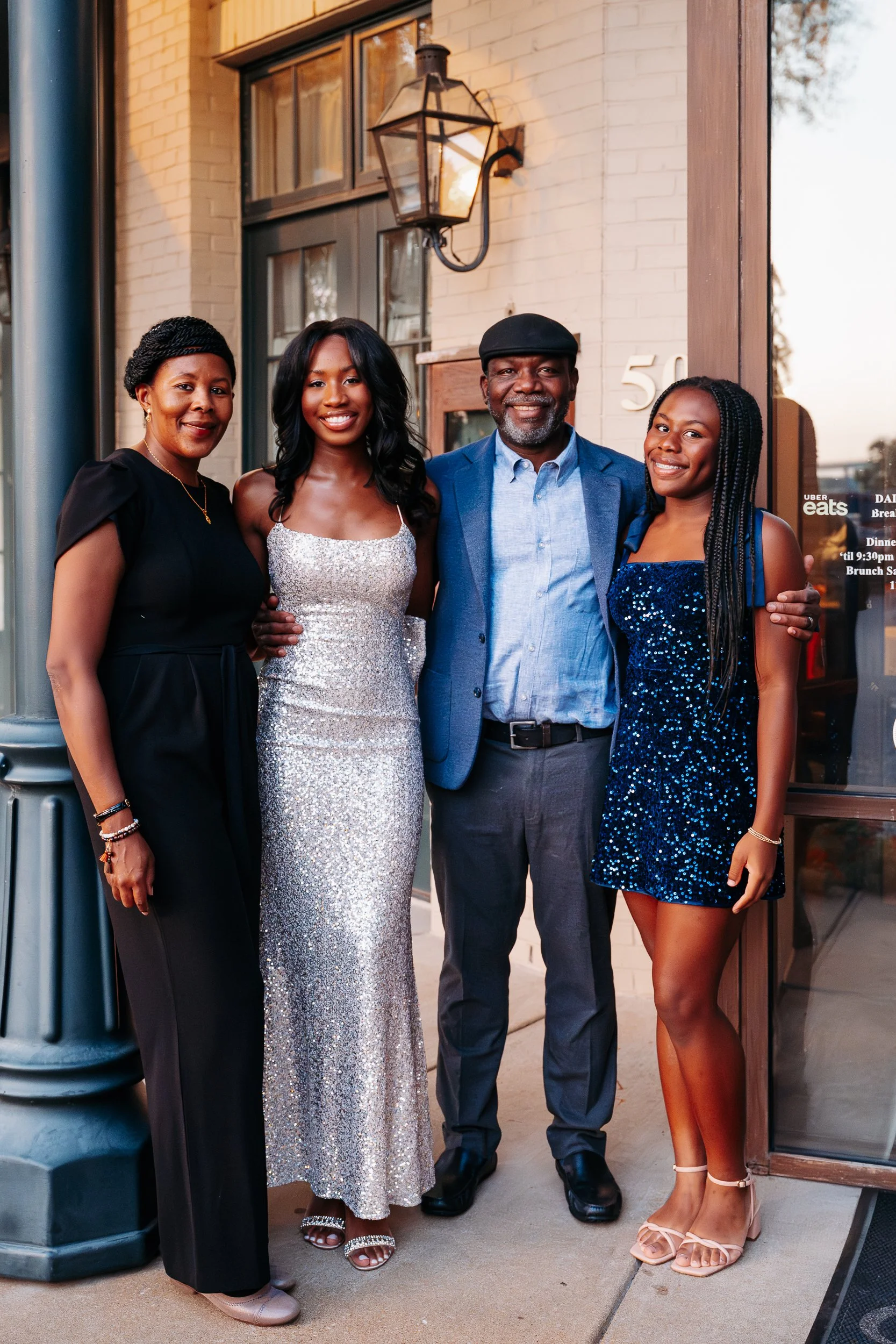 A group of five people, two women and three men, standing outside a building with a light brick wall and a window, posing for a photo. They are dressed in evening attire, with all smiling and close together.