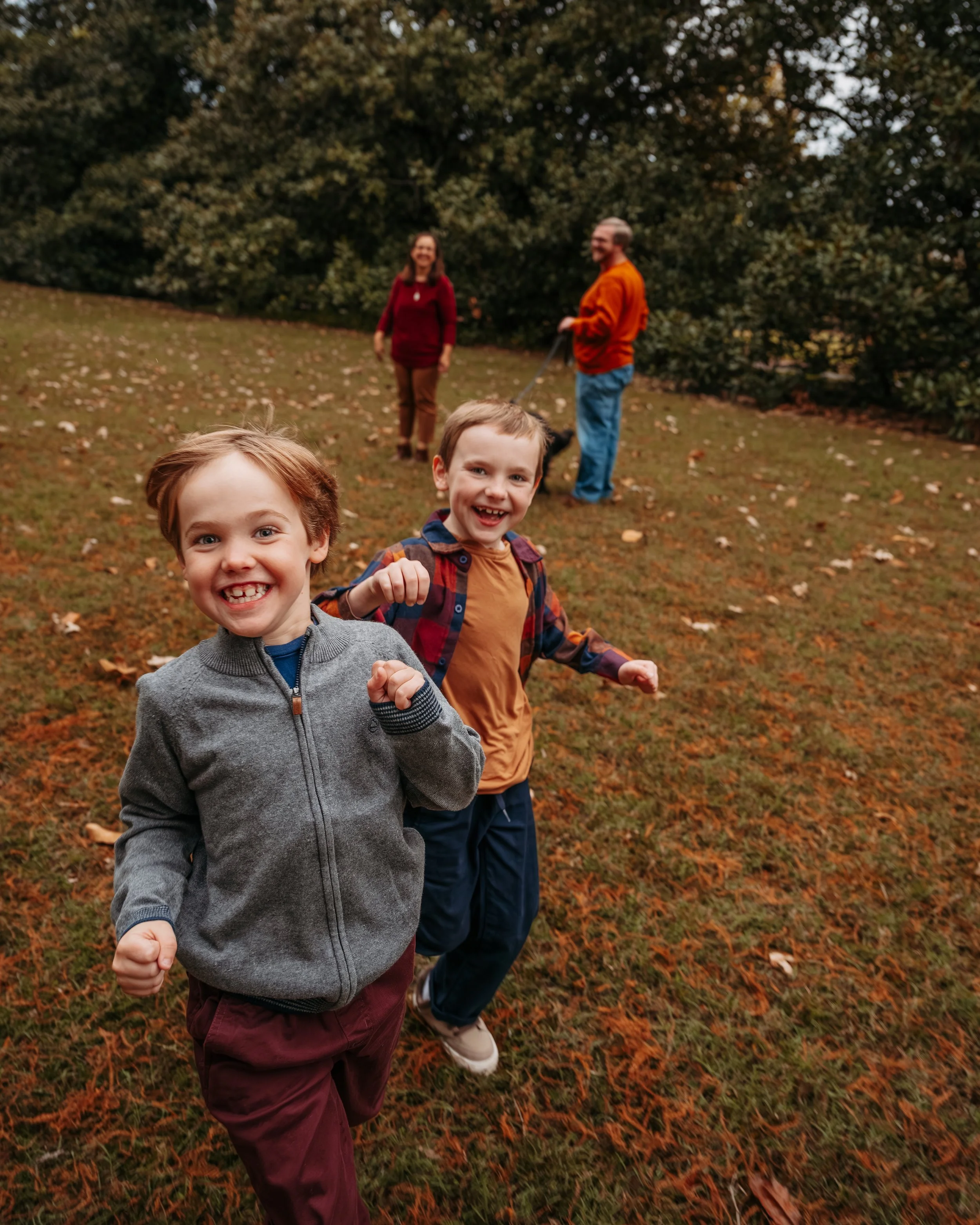 Two young boys smiling and running outdoors on a fall day, with a woman and a man walking a dog in the background under trees with falling leaves.