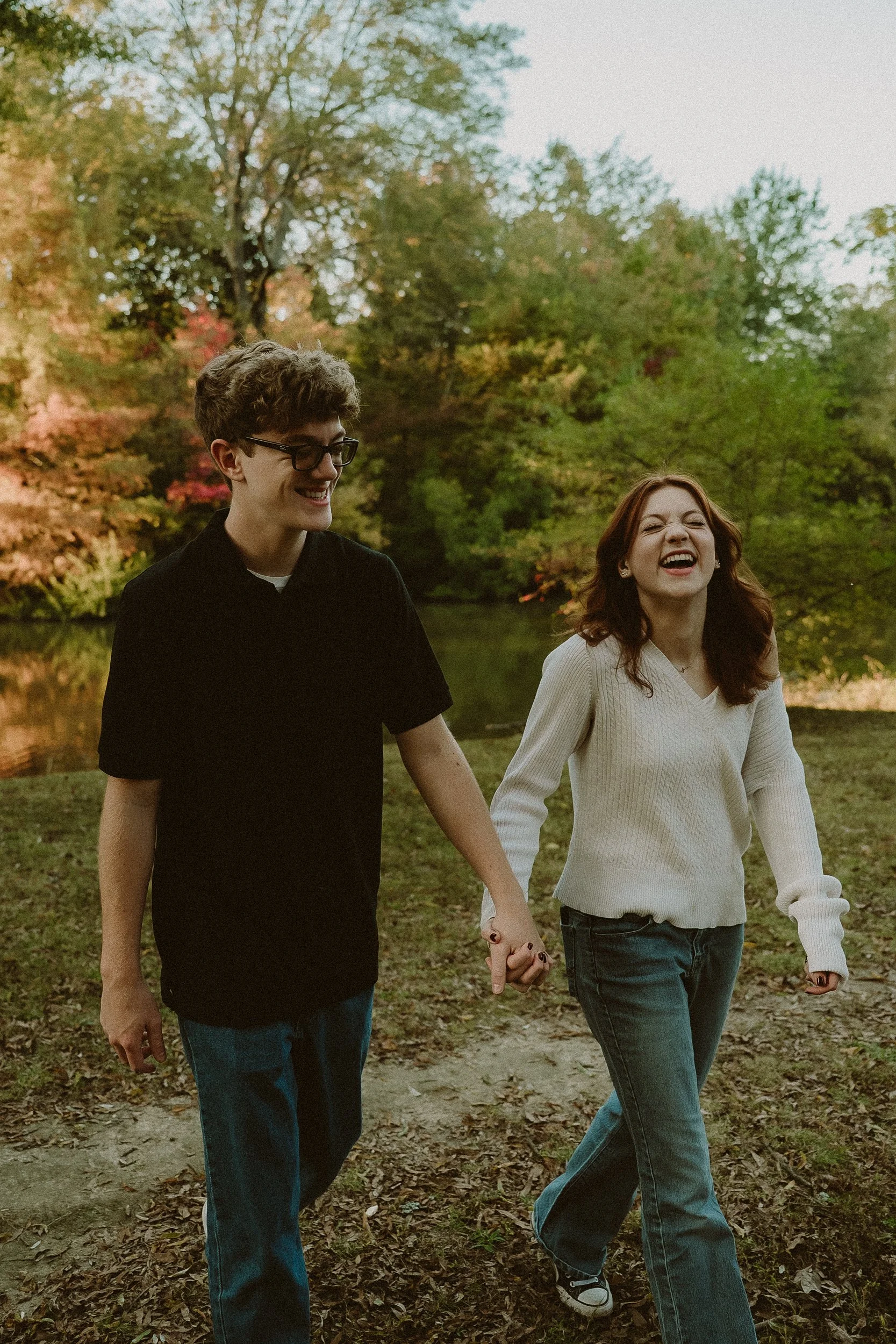 A young man and woman holding hands and walking outdoors in a park during autumn, smiling and laughing.