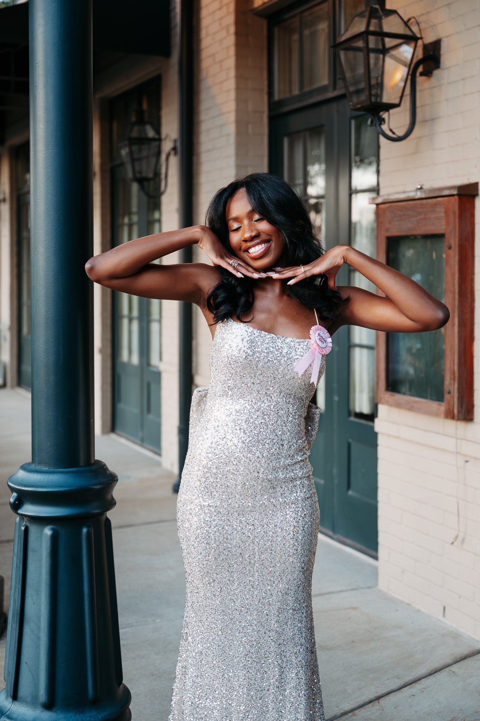 A woman in a silver, sparkly dress with a pink ribbon and badge smiling with eyes closed, standing outside a building with large windows, door, and outdoor lanterns.
