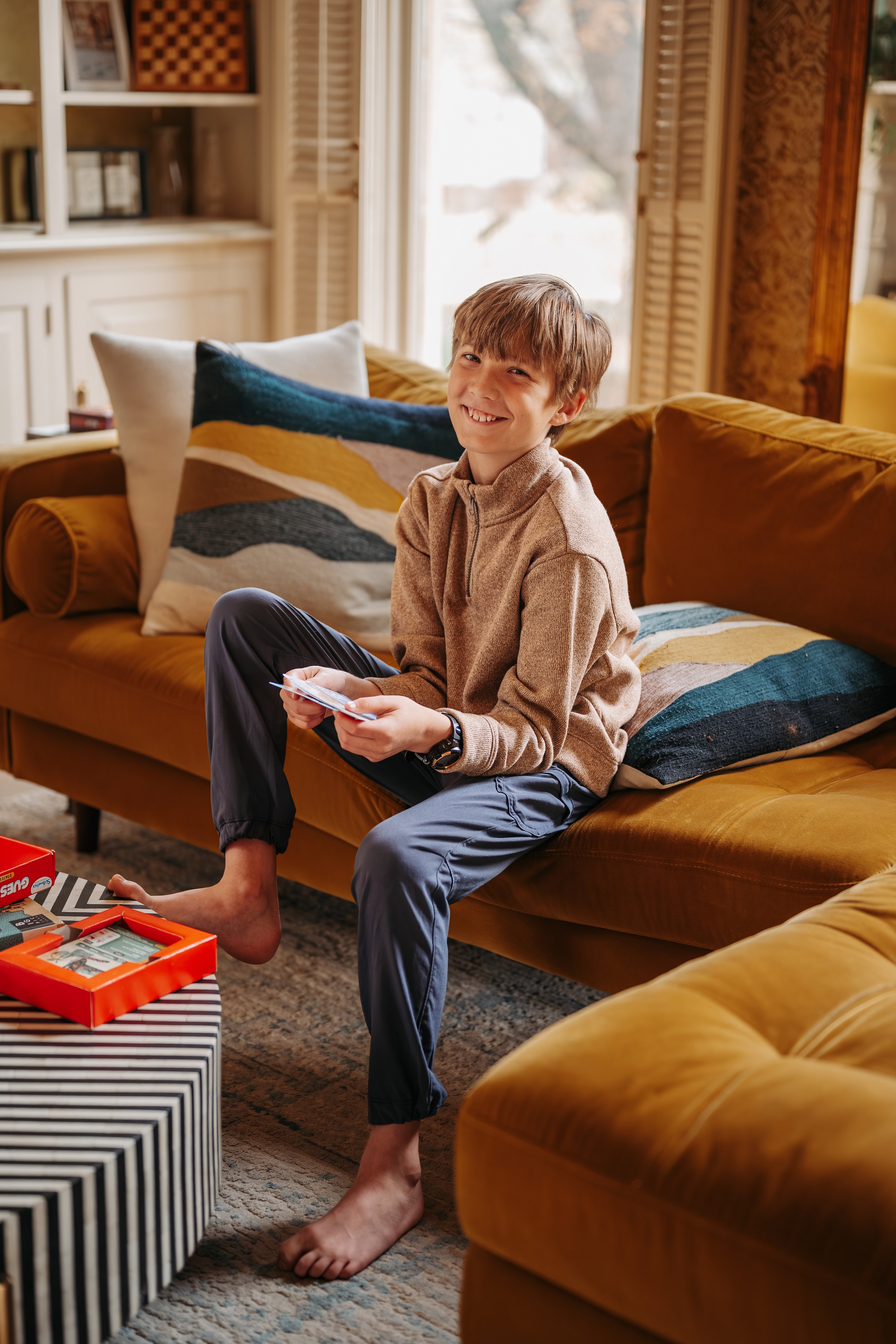 A boy sitting on a mustard-colored sofa, smiling and holding a card, in a cozy living room with patterned pillows and a window in the background.