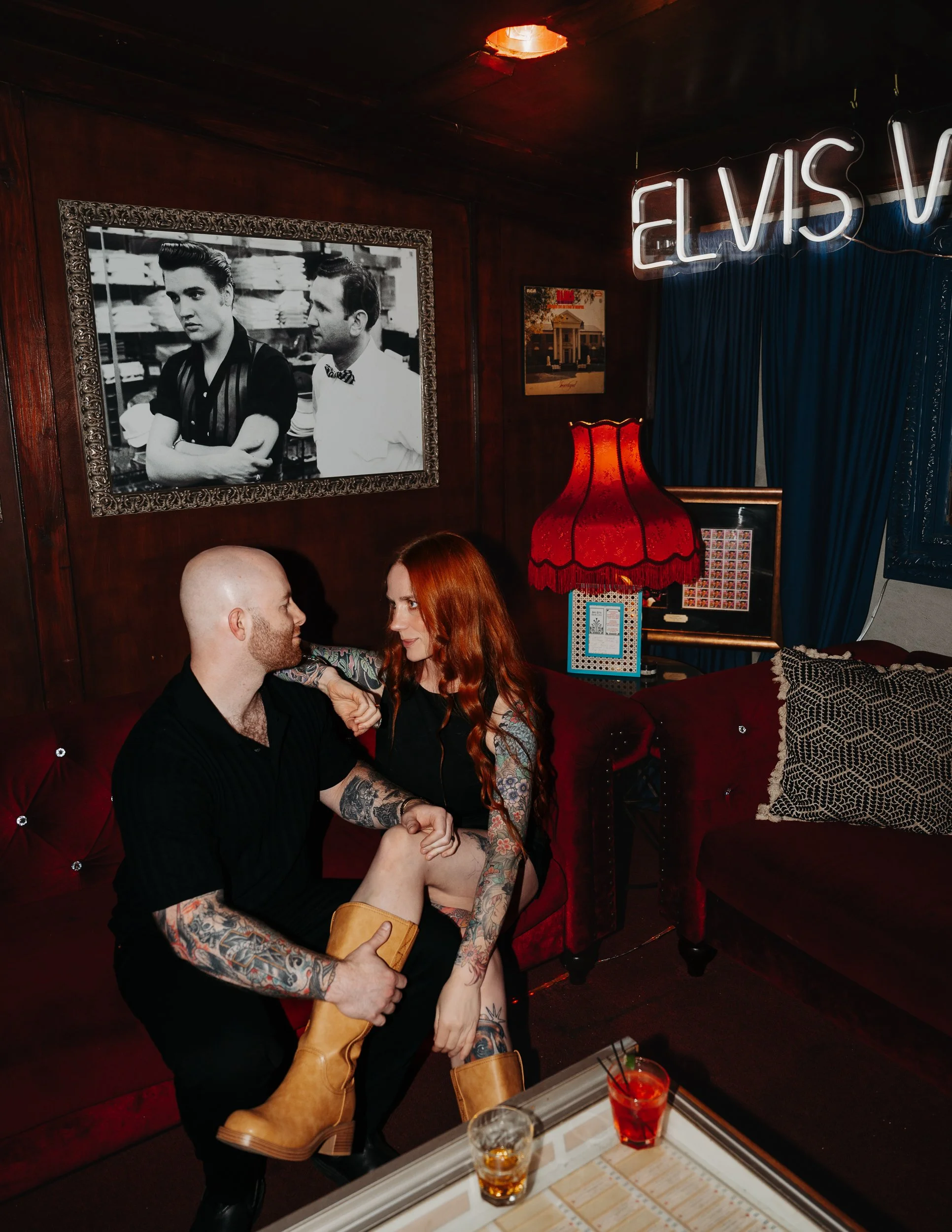 A man and woman with tattoos sitting on a red velvet couch in a dimly lit room. The man is bald and has tattoos on his arms, while the woman has long red hair and tattoos on her arms and legs. They are facing each other closely. There are drinks on a table in front of them. The room has dark wood walls, a large black-and-white framed photo, a red lamp, a blue curtain, and a neon sign that reads 'ELVIS V'.