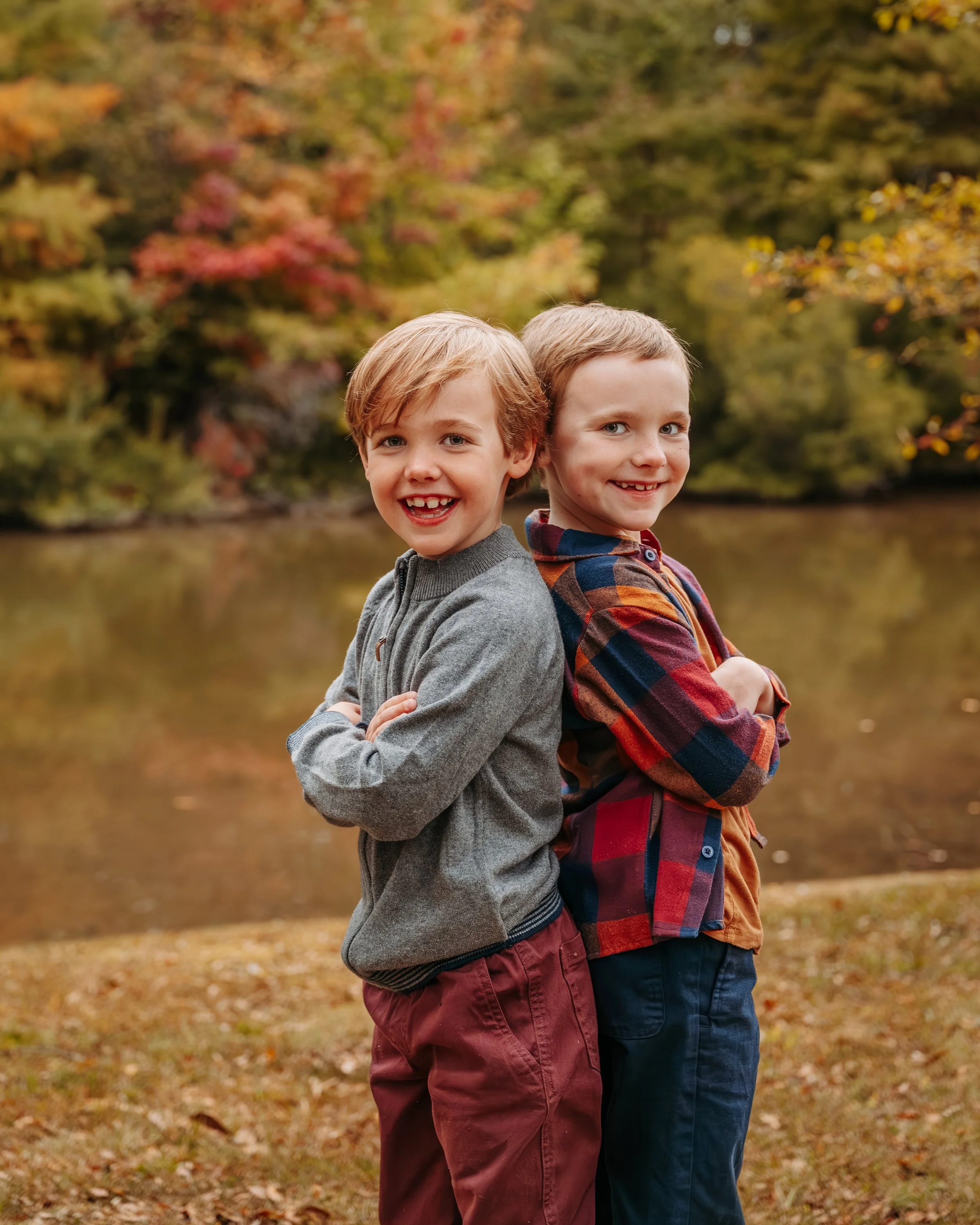 Two young boys standing back to back by a river in autumn, smiling with folded arms, surrounded by fall foliage.