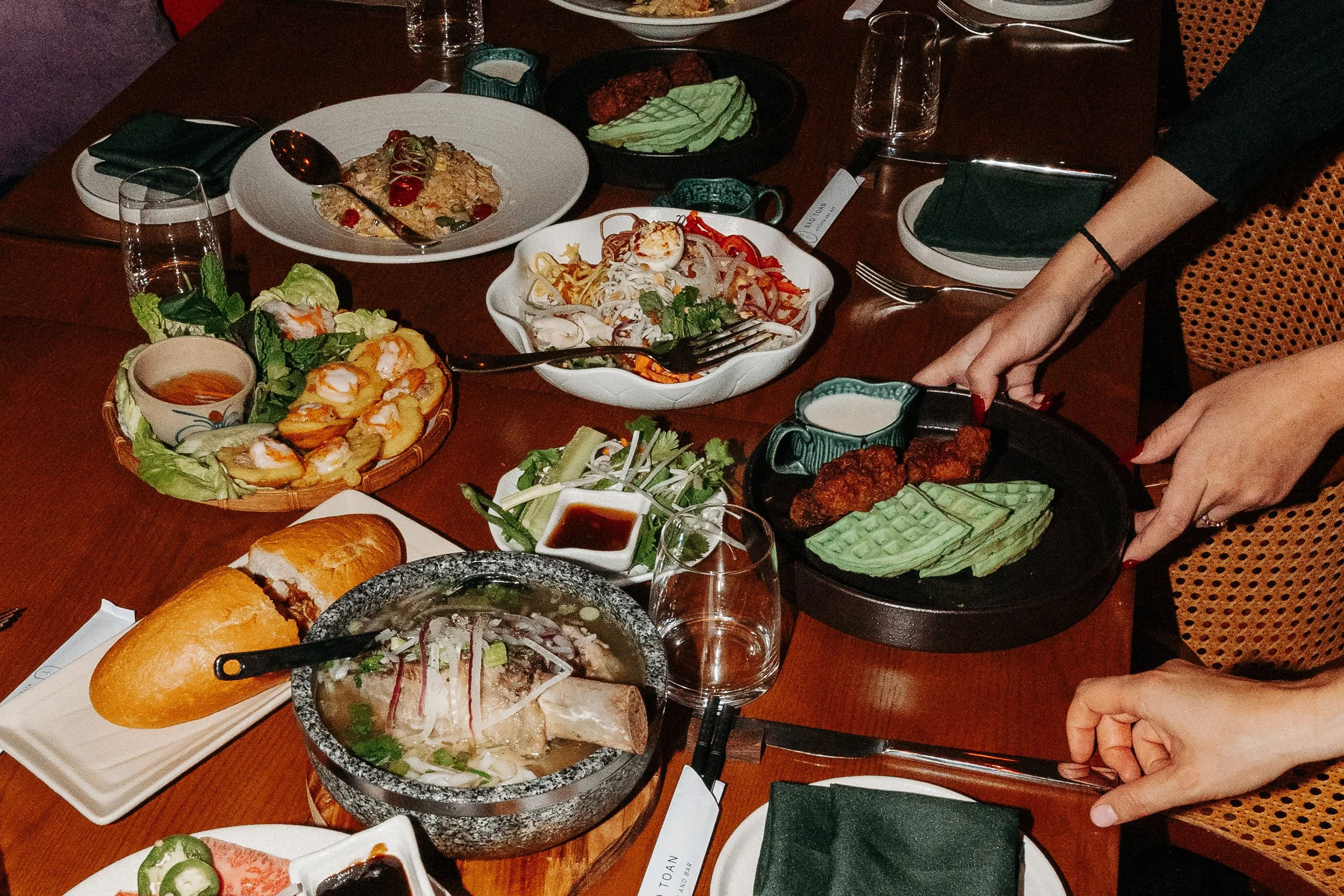 A table with various Asian dishes, including a bowl of ramen, spring rolls, and Asian-style fried chicken, along with vegetables and dipping sauces, with hands placing a dish on the table.