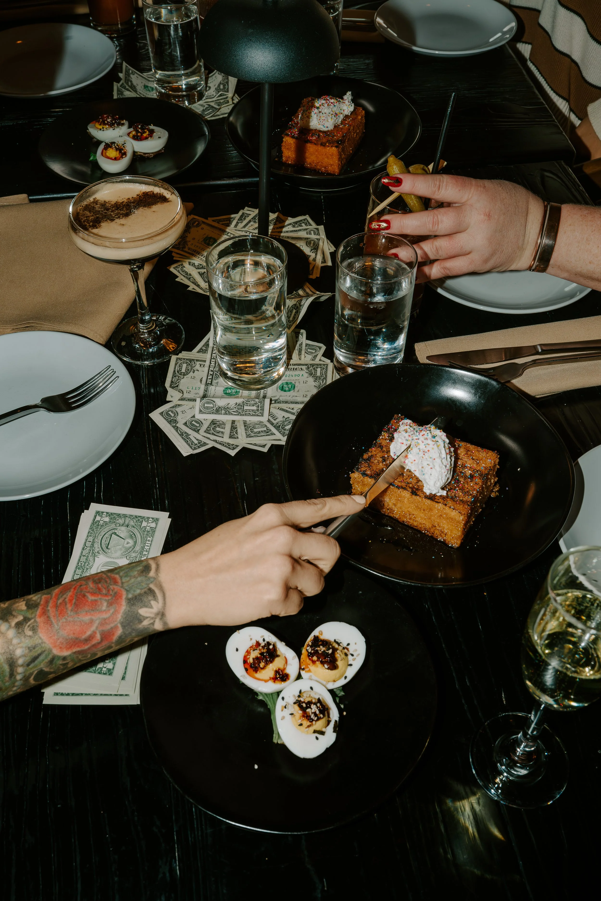 A table with various desserts, drinks, and scattered dollar bills. There are small deviled eggs, a slice of cake with ice cream, a creamy dessert, and several glasses of water and a cocktail. A person's hand with a tattoo and painted nails is cutting