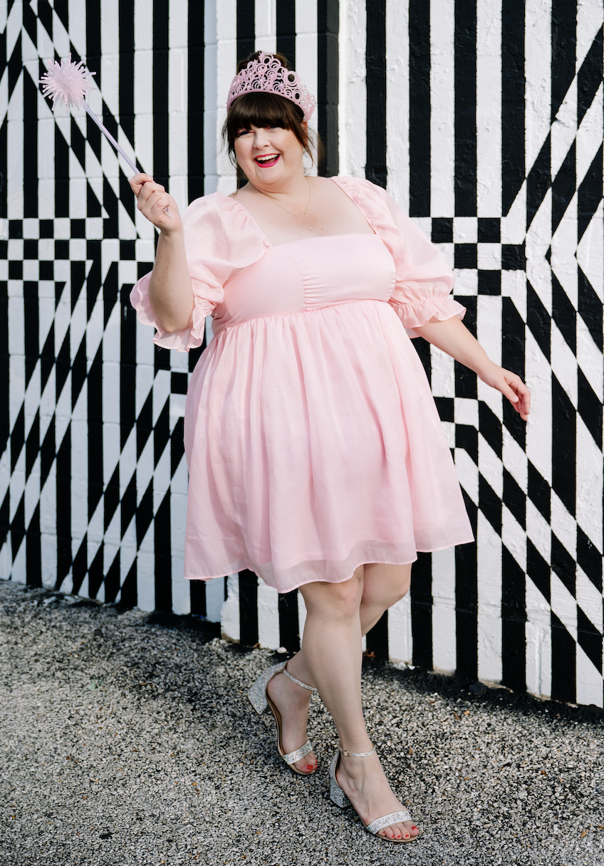 A woman dressed as a princess wearing a pink dress, pink tiara, and holding a pink wand with a star, standing in front of a black and white geometric patterned wall.