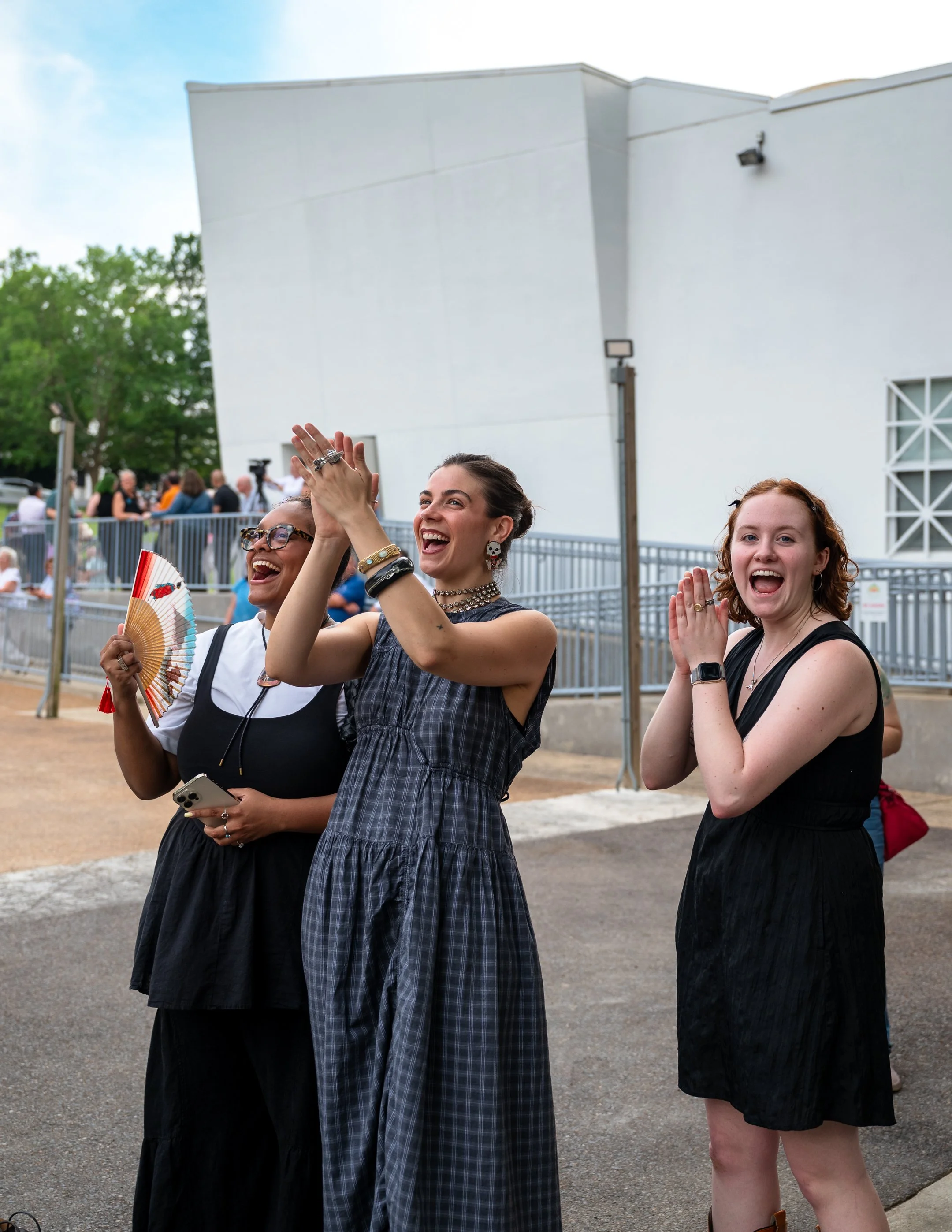 Three women laughing and celebrating outdoors at a public gathering or event, standing in front of a white building.