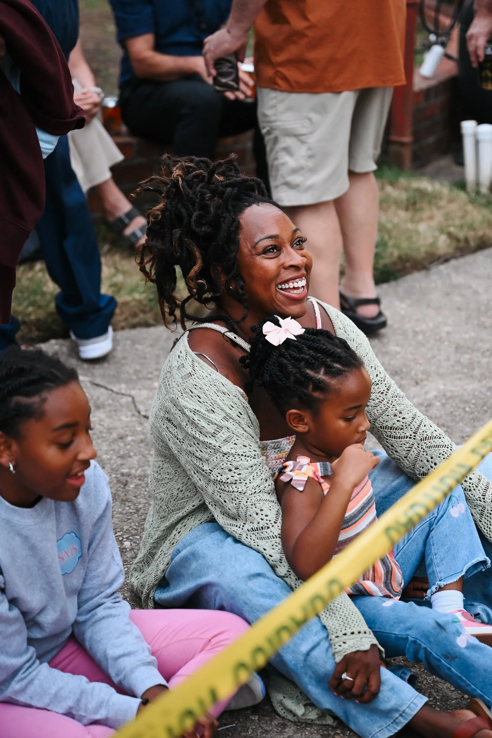 Smiling woman and two young girls sitting on the ground outdoors, surrounded by a crowd, with yellow caution tape in front.