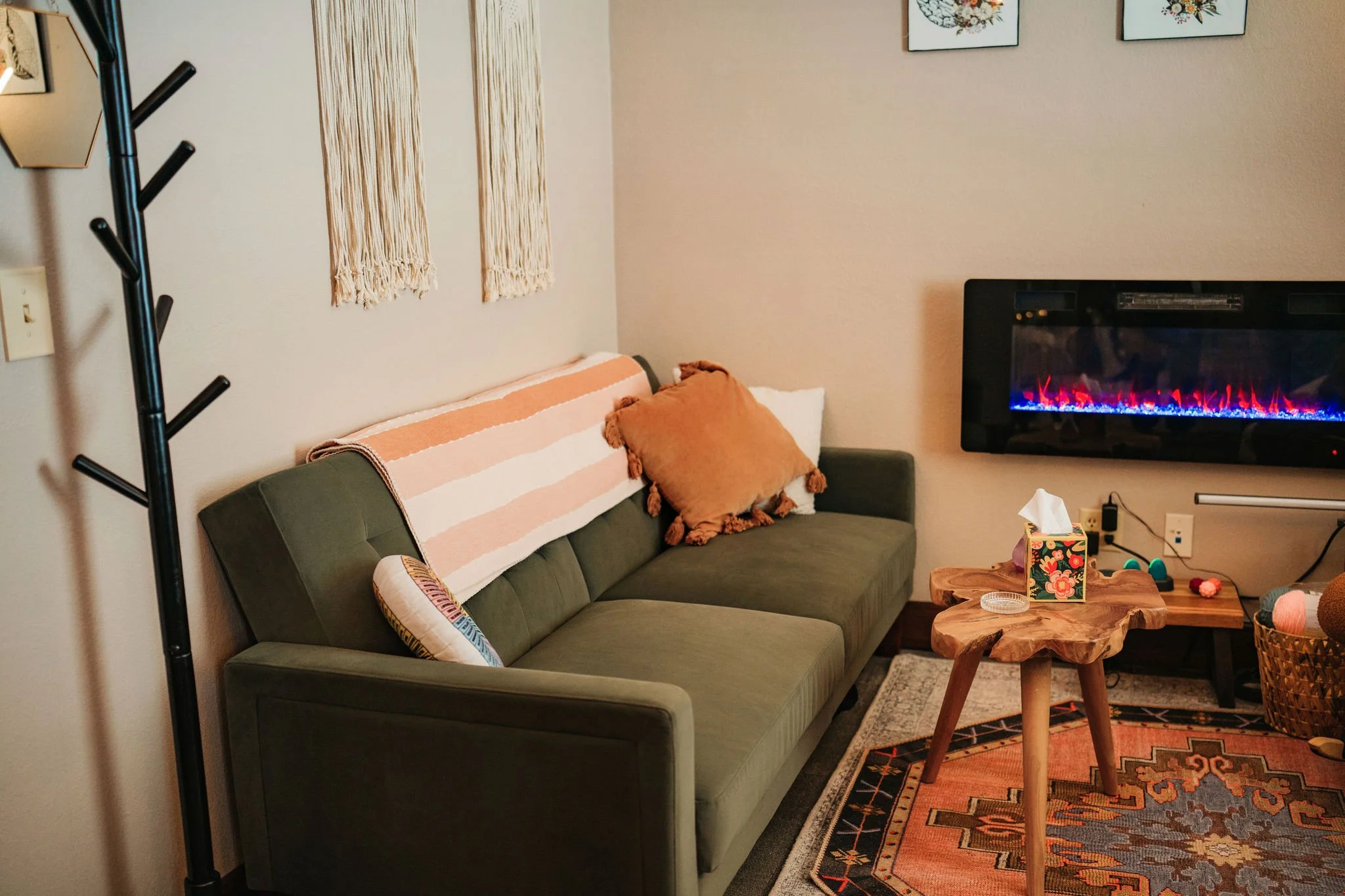 Counseling office with a green sofa decorated with pillows and a striped blanket, a wooden coffee table, a wall-mounted electric fireplace, and wall art. There is a side basket with yarn and a tissue box on the coffee table.
