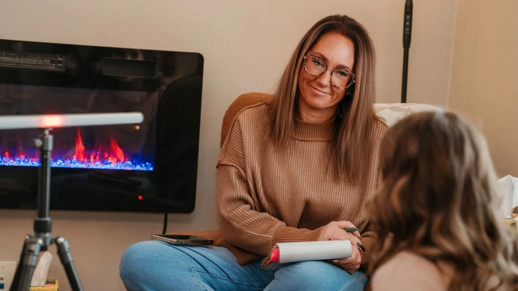 A woman with long brown hair, glasses, and a tan sweater sitting on a beige chair, smiling while talking to a young girl with wavy brown hair. A small electric fireplace with blue and red flames is visible in the background.