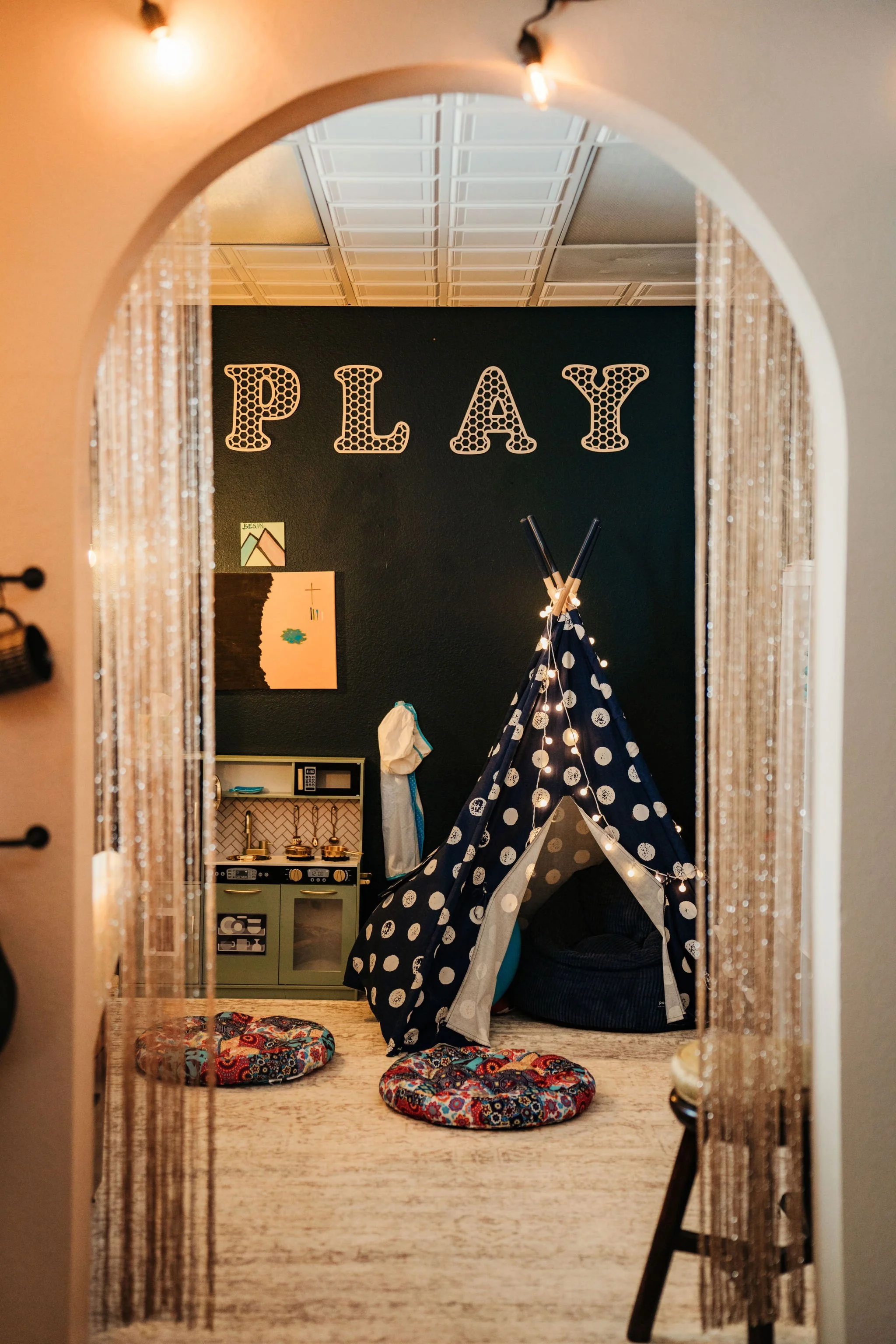 Children's playroom featuring a dark wall with the word 'PLAY' in large decorative letters, a polka dot teepee tent with string lights, two colorful circular cushions, a small play kitchen, and children's artwork on the wall.
