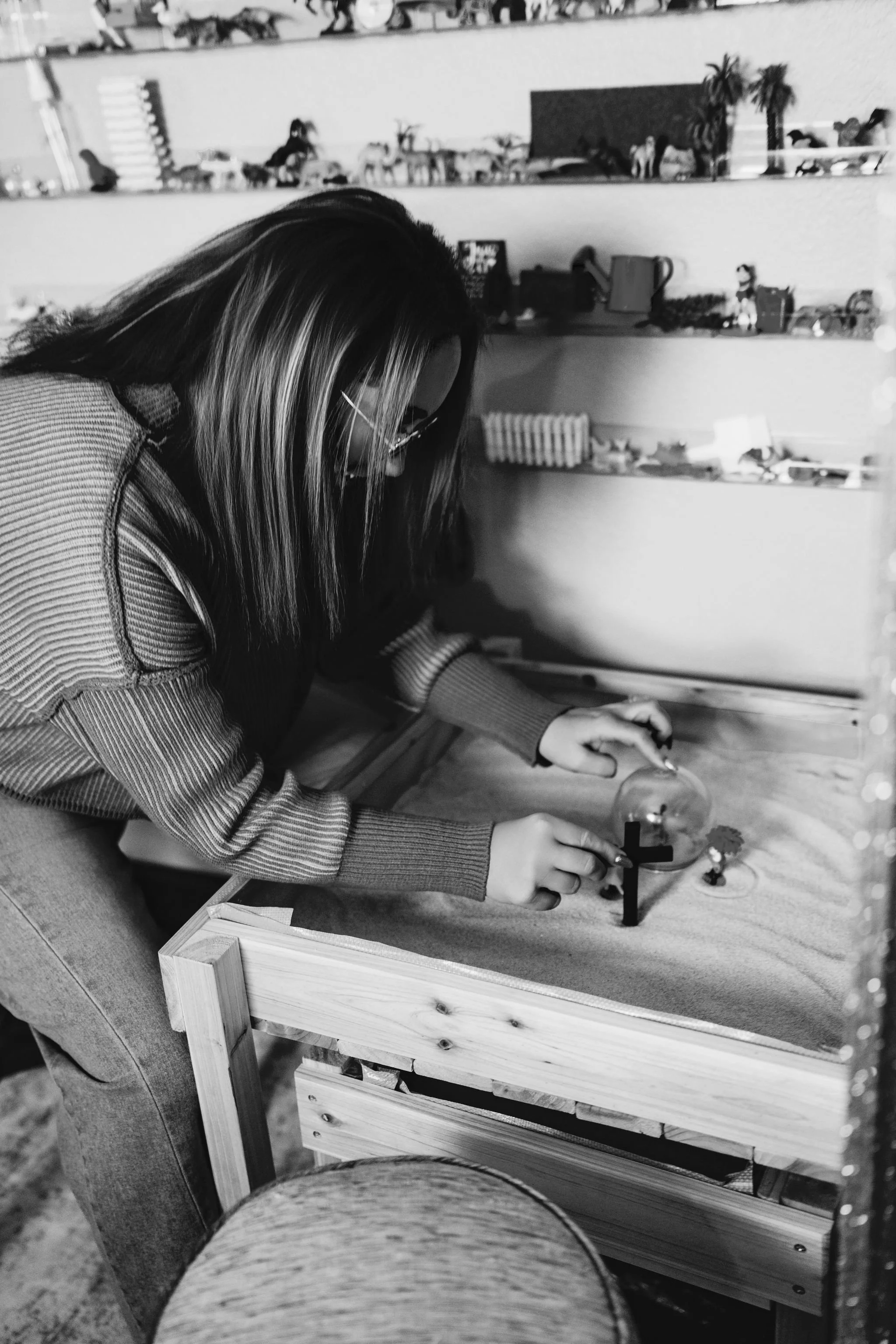 A woman playing a tabletop game with a glass globe and miniature figures on a grass-textured surface, in a room with shelves filled with toys and decorations.