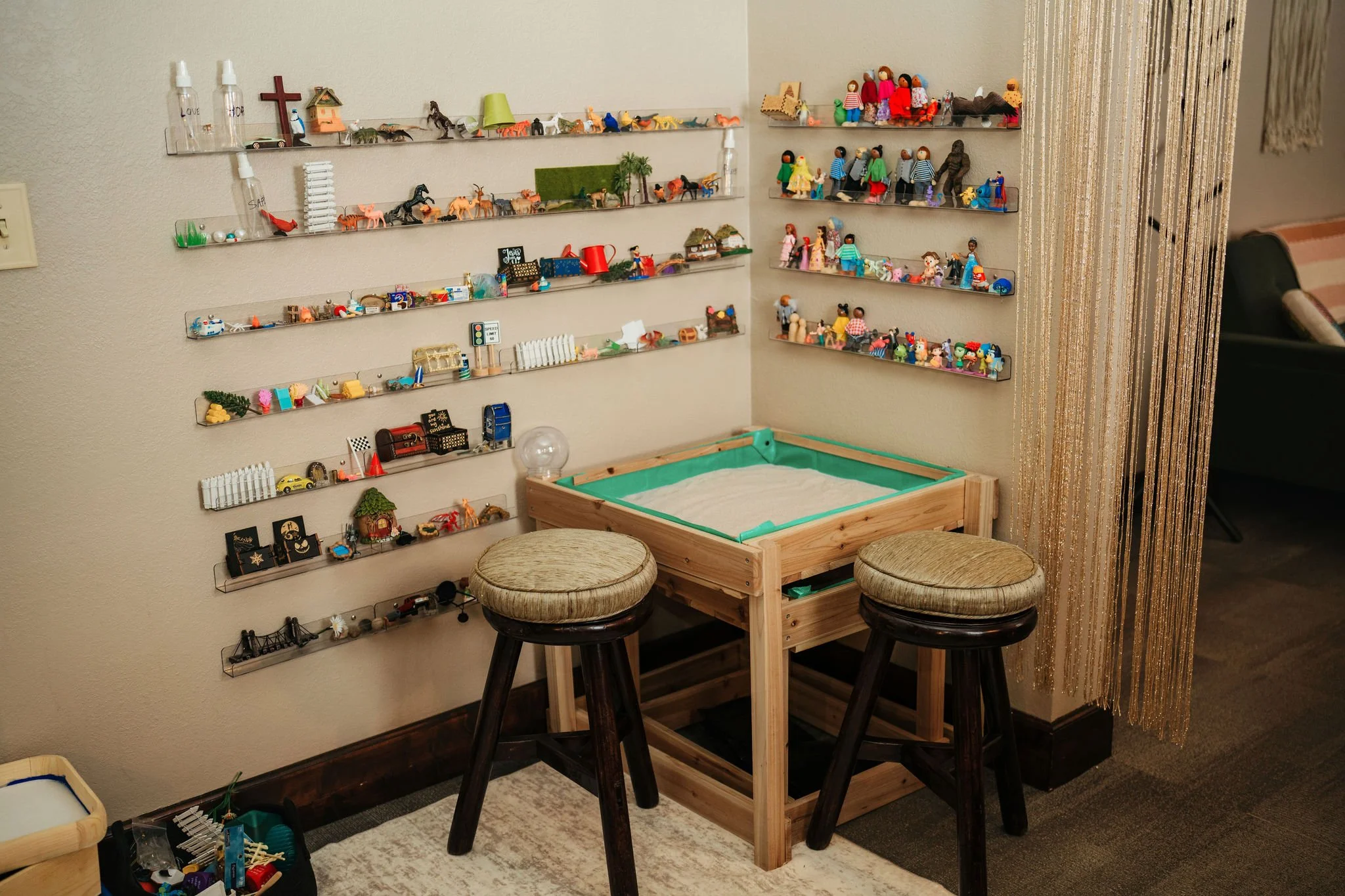 A corner of a room with two wooden stools and a rectangular sandbox. The wall has multiple clear shelves filled with miniature toys, figurines, and small decorations. To the right is a gold, beaded curtain.