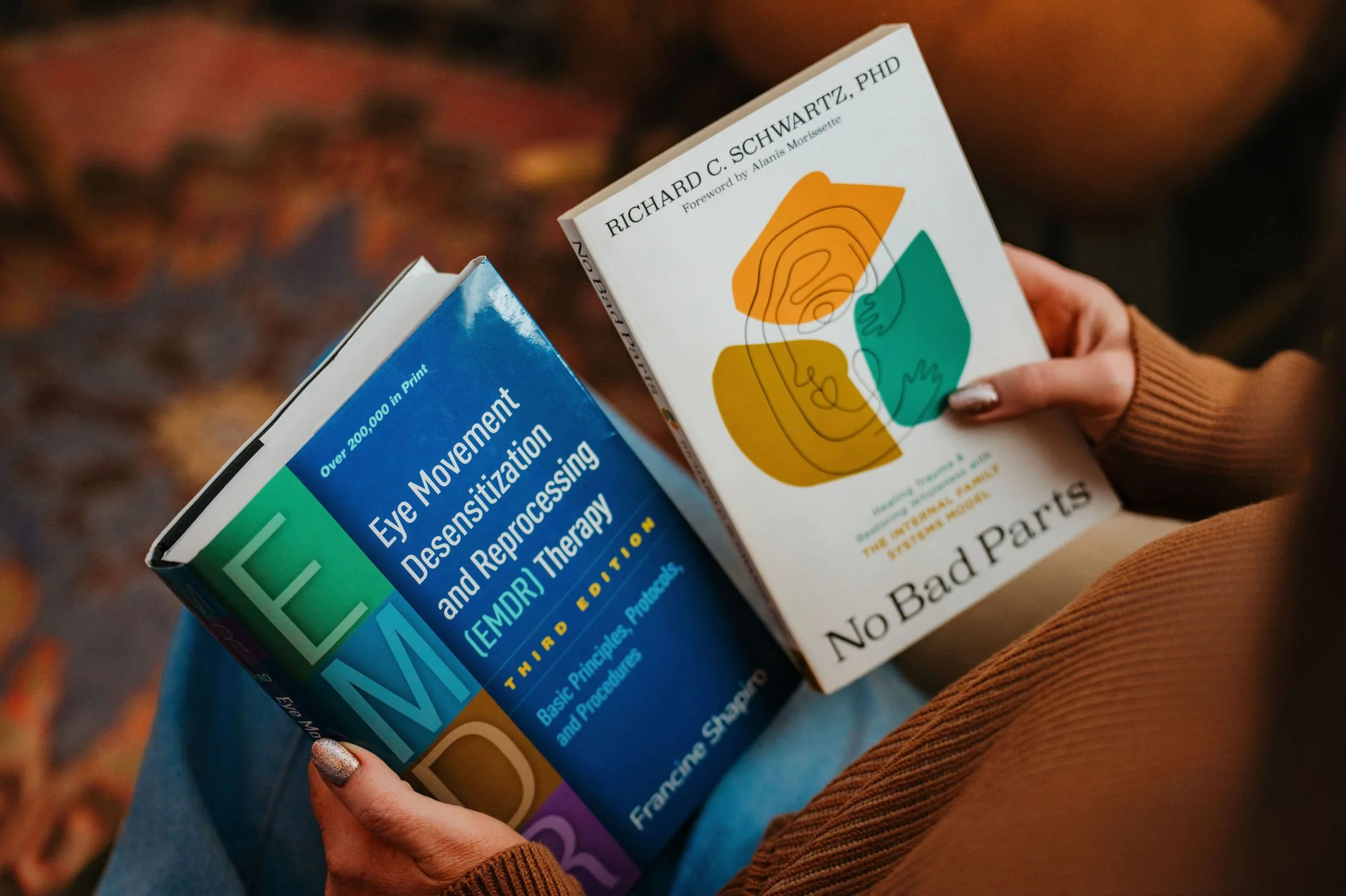A person sitting on a chair or sofa holding two books about eye health and therapy, one titled 'Eye Movement Desensitization and Reprocessing (EMDR) Therapy' and the other 'No Bad Parts' by Richard C. Schwartz, PhD. The person is wearing a brown sweater and has silver, glittery nail polish.