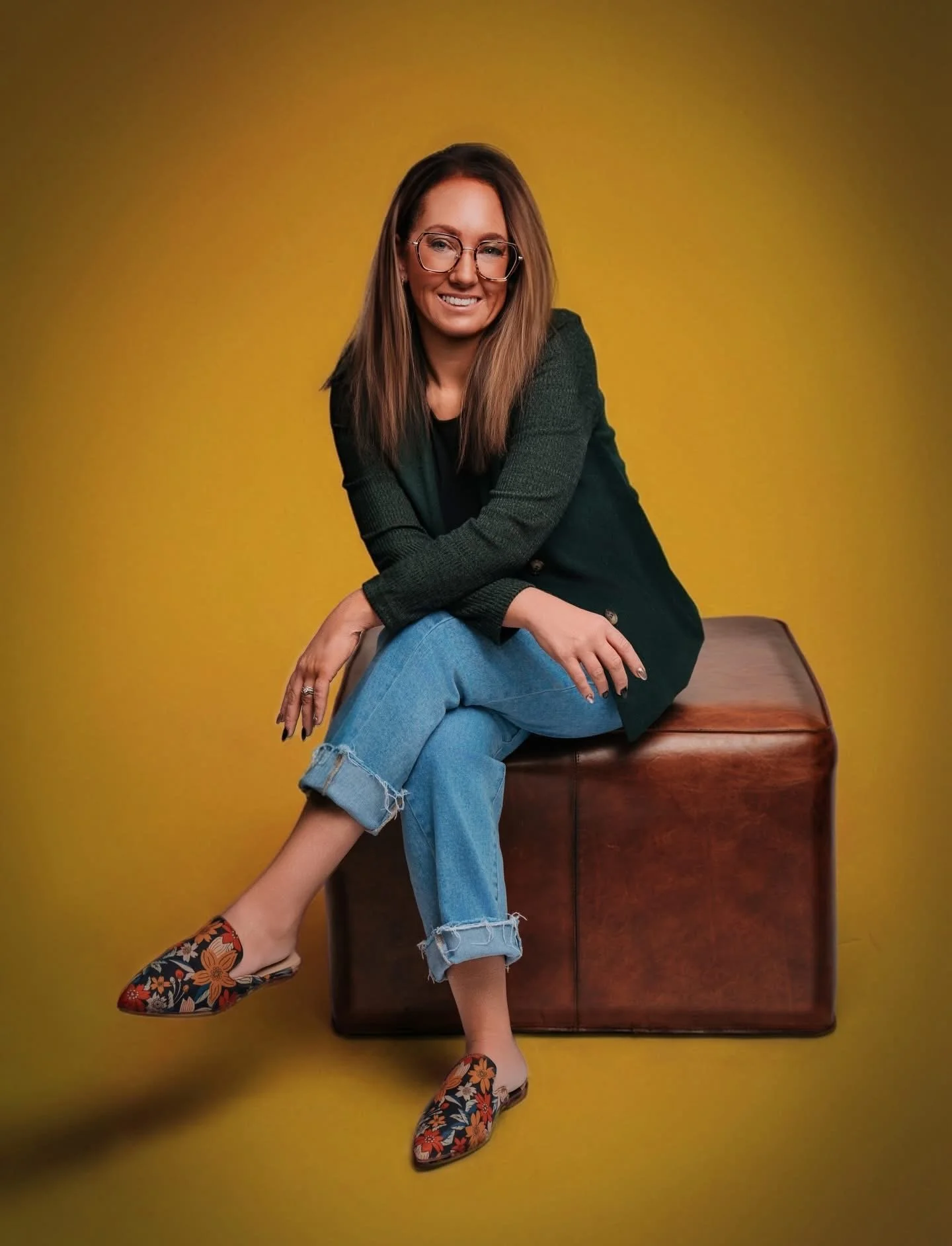 Woman sitting on a brown leather ottoman against a yellow background, smiling, wearing glasses, a dark blazer, ripped jeans, and floral-patterned slip-on shoes.