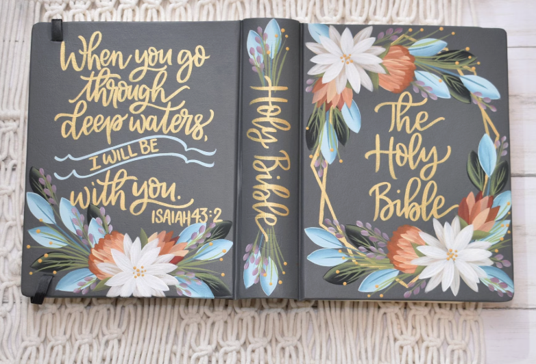 Open Bible with floral decorations and hand-lettered text reading, 'When you go through deep waters, I will be with you. Isaiah 43:2' on the left page and 'The Holy Bible' on the right page.