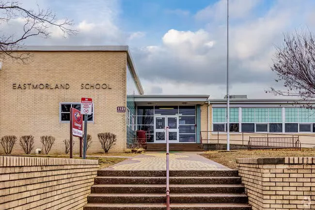 Front entrance of Eastmorland School building with brick stairs, a ramp, and a flagpole, under a partly cloudy sky.