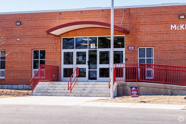 Exterior of a brick building with glass doors, multiple windows, red metal handrails, and a ramp. There's a sign near the ramp and the address 610 is visible on the building.