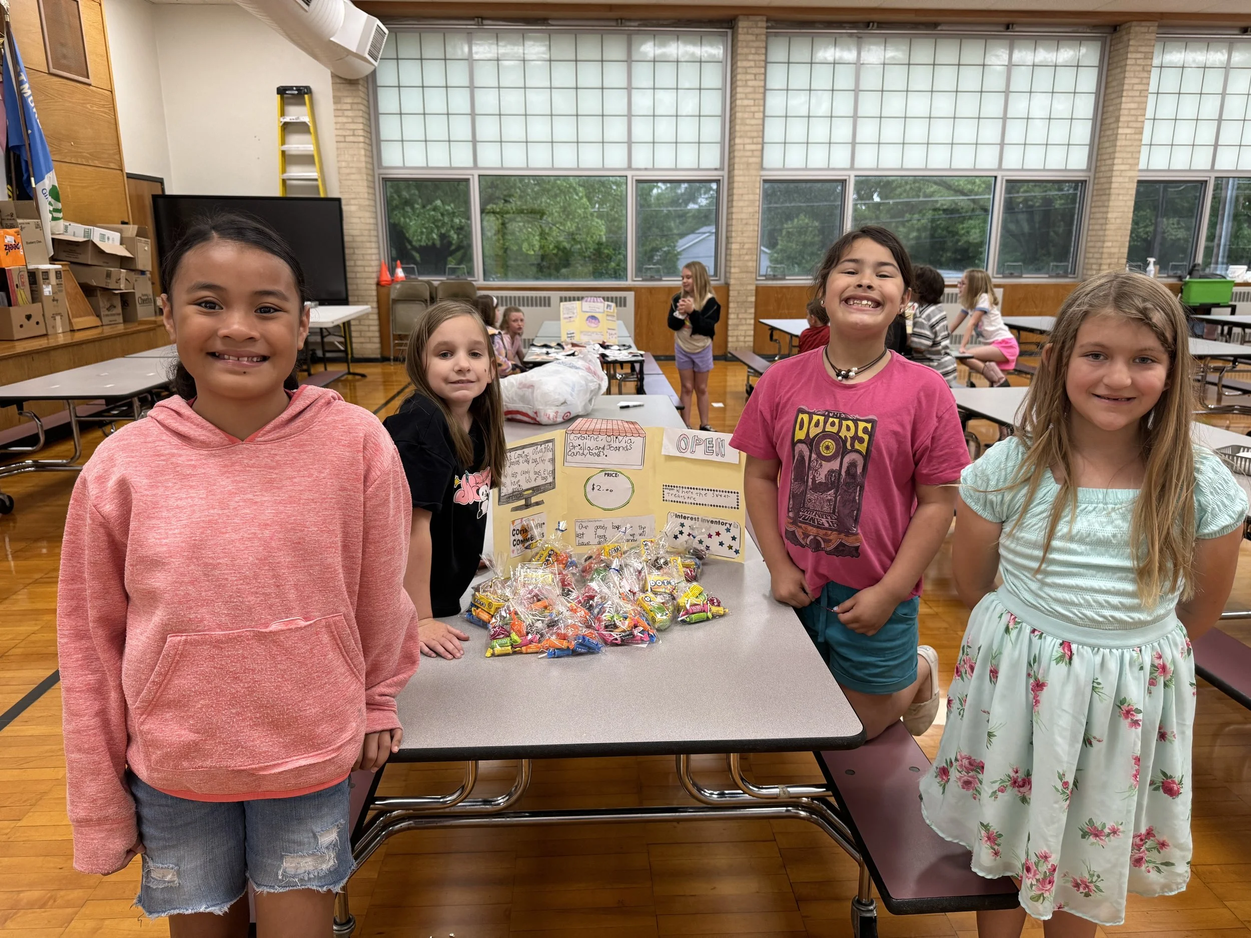 Four young girls standing in front of a display table with a variety of candies, inside a school cafeteria or multipurpose room with large windows and tables.