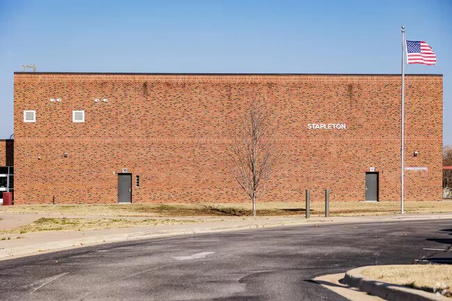 Brick building with the word 'Stapleton' on it, two doors, a leafless tree, a flagpole with the American flag, and a clear blue sky.