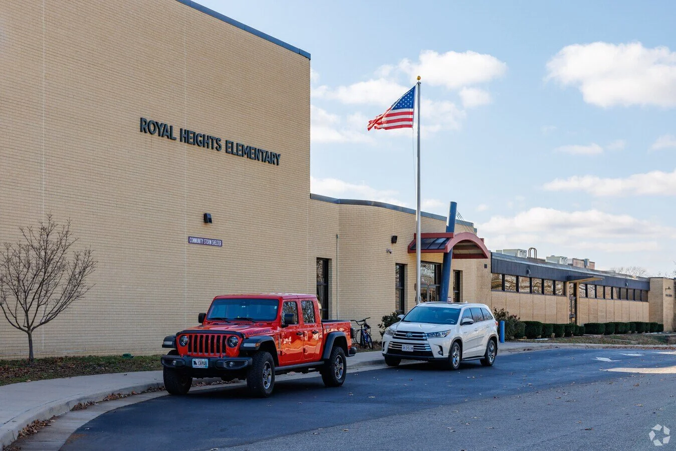Exterior view of Royal Heights Elementary school with a red Jeep, white SUV parked in front, American flag on pole, and clear sky.