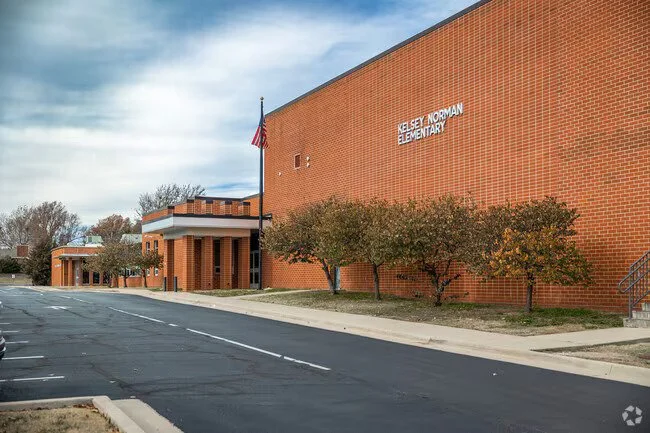 Exterior view of a Kelsey Norman elementary school building with a sign that reads 'Kelsey Norman Elementary' and a parking lot in front.